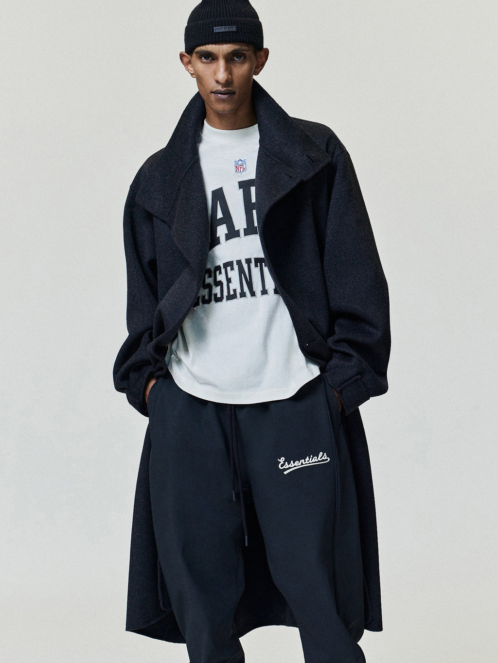 NFL Paris Merch Tee in off white with bold black Paris Essentials lettering layered under a black coat and styled with dark relaxed trousers Tops