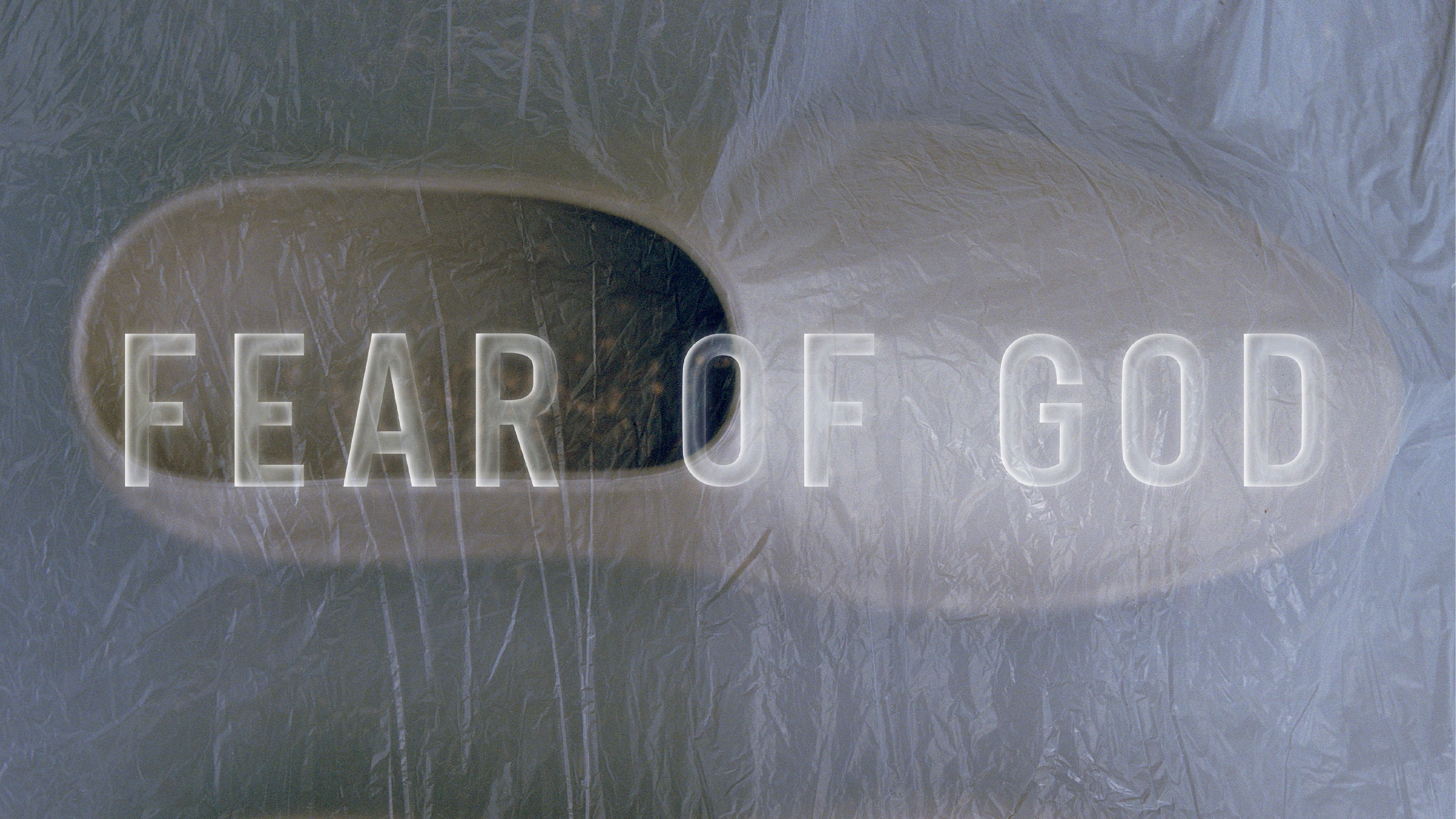 Abstract overhead view of two smooth, rounded stone objects placed side by side on a textured surface, with the words "FEAR OF GOD" softly illuminated across them in a modern, translucent font.