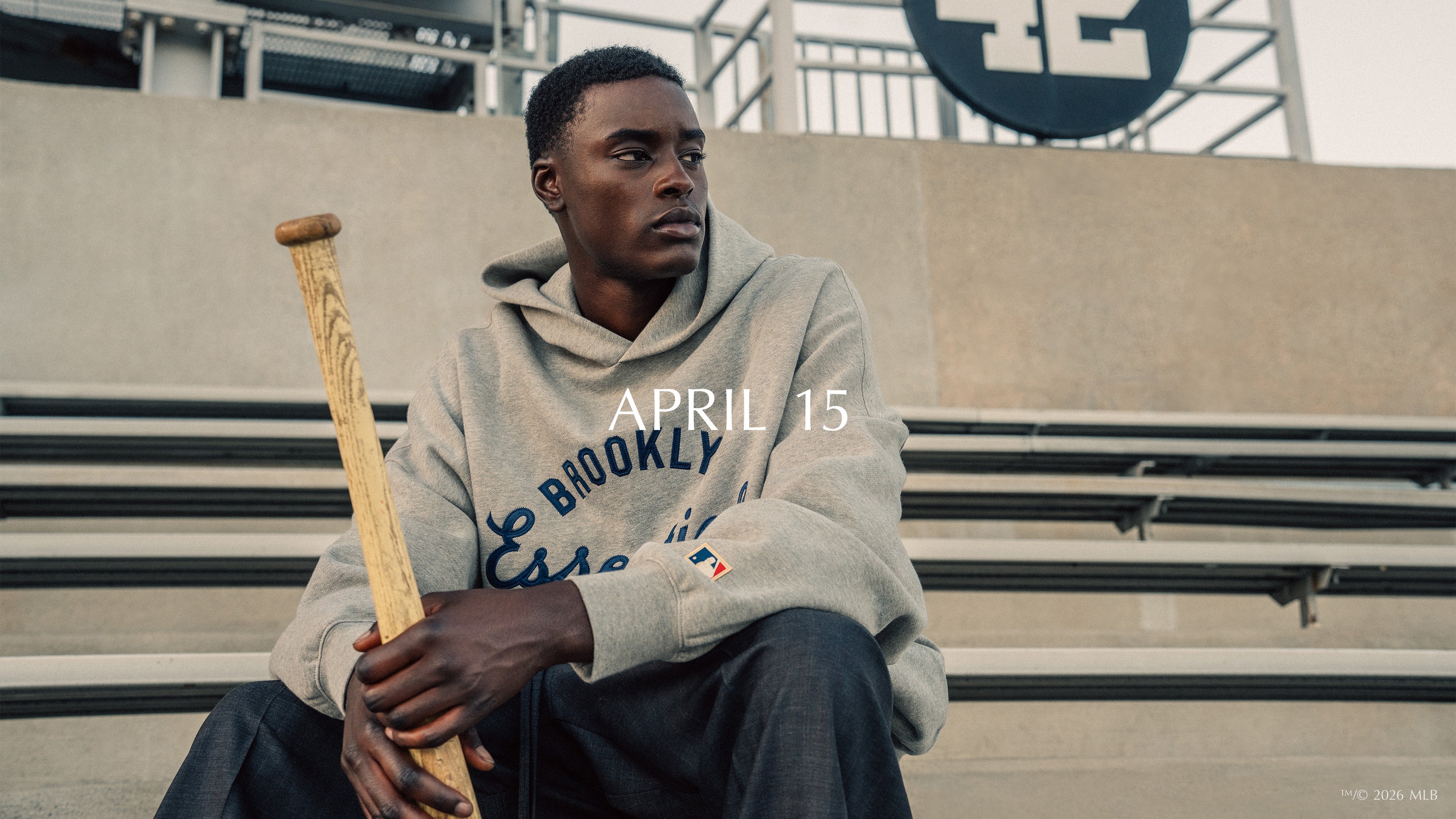 Man in gray Brooklyn Essentials hoodie sitting on stadium bleachers holding wooden baseball bat looking away