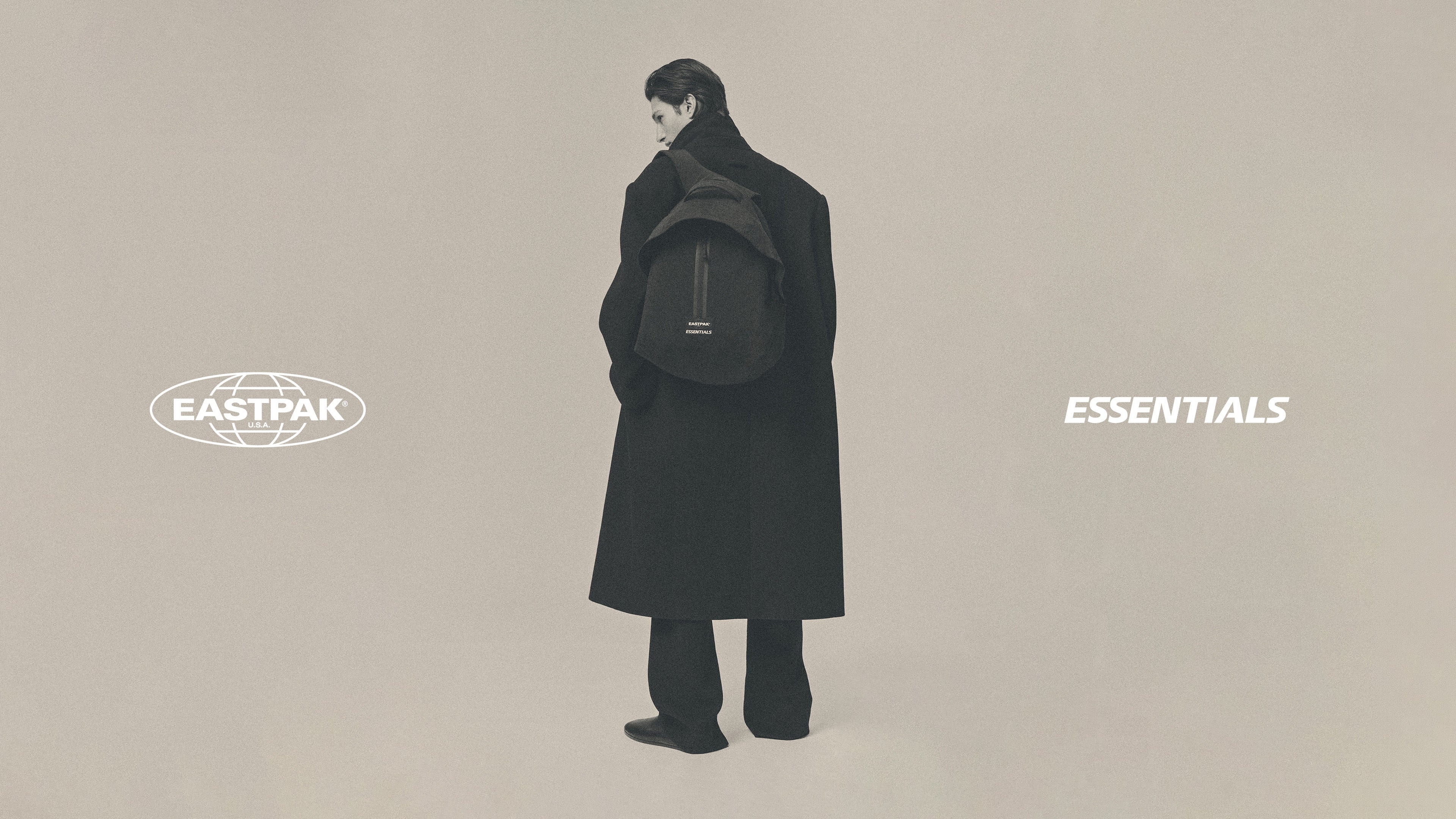 Person in a long black coat wearing a black backpack and standing against a muted beige background with Eastpak and Essentials logos nearby