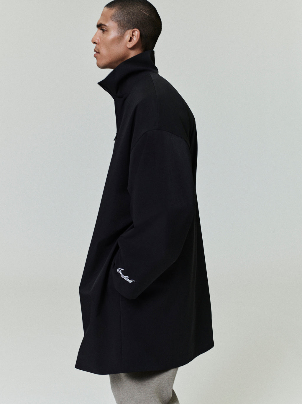 Side view of man wearing black sporty nylon high neck jacket with embroidered detail on sleeve against plain background
