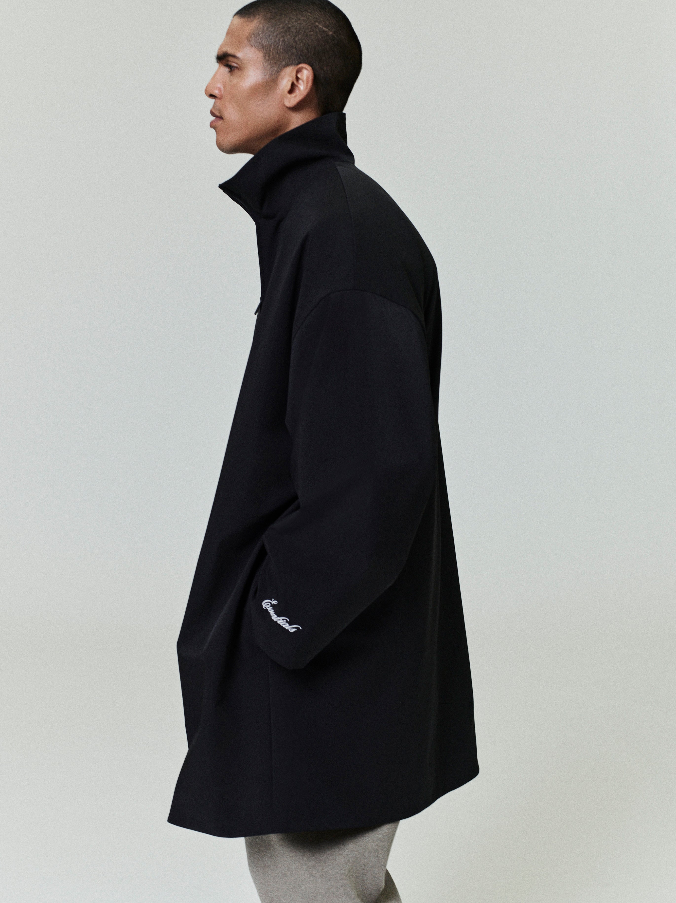 Side view of man wearing black sporty nylon high neck jacket with embroidered detail on sleeve against plain background