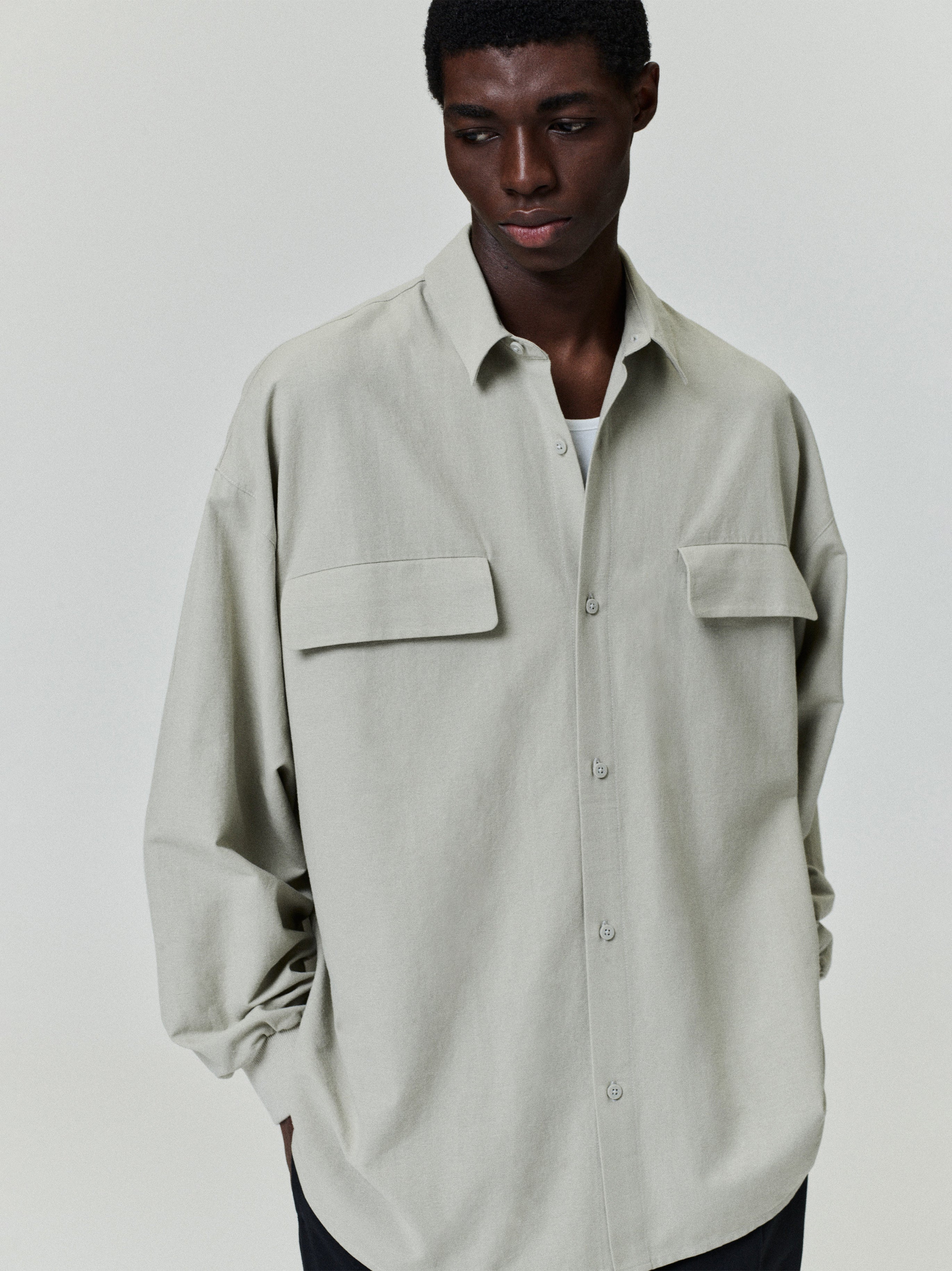 male model wearing oversized light beige Oxford shirt with long sleeves and flap pockets against plain background