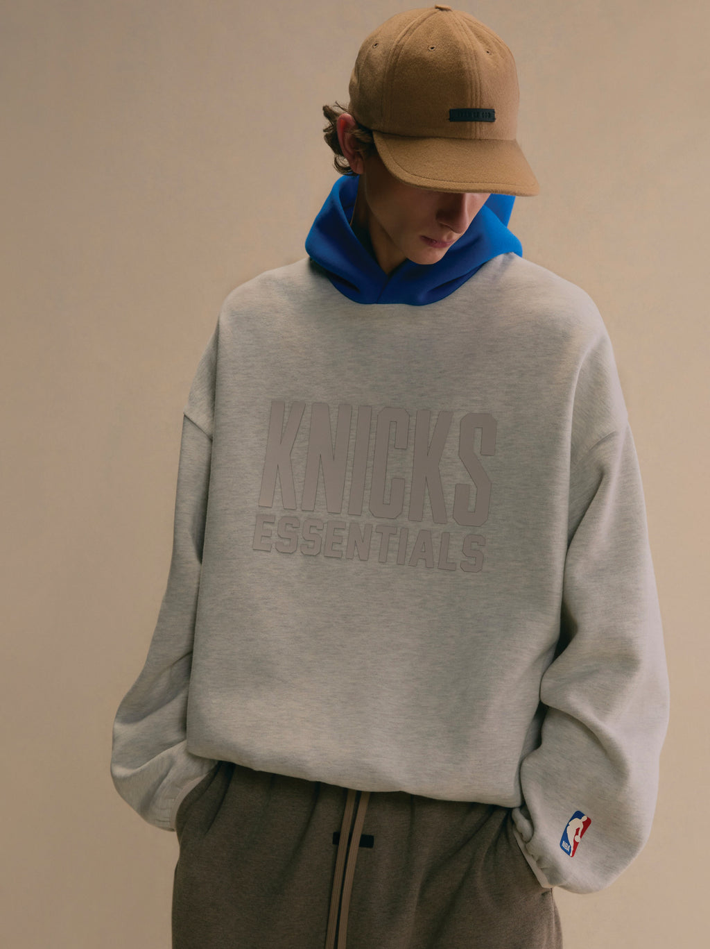 Young man wearing light grey Knicks Essentials hoodie with subtle logo and blue hood layered underneath against plain background