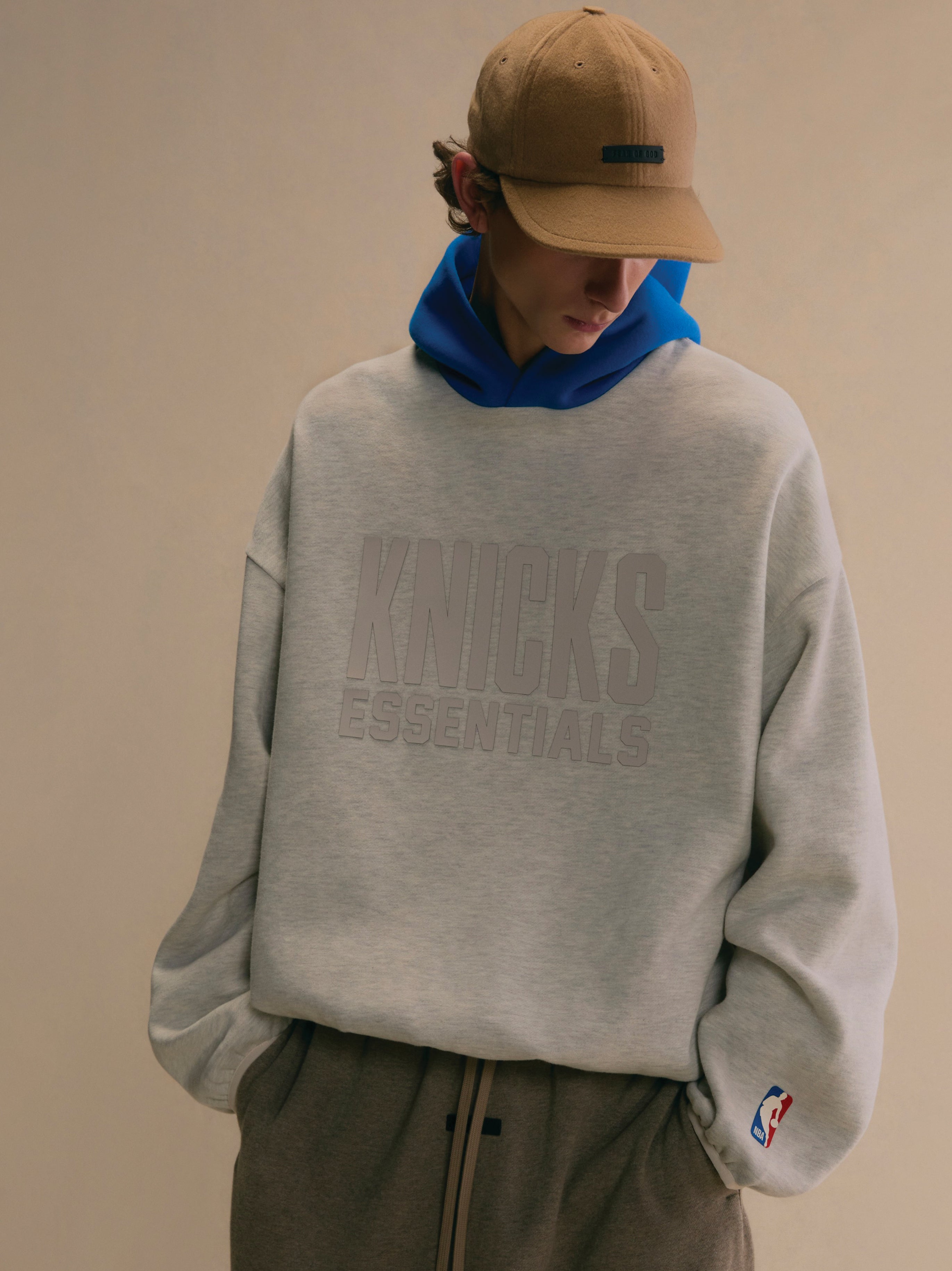 Young man wearing light grey Knicks Essentials hoodie with subtle logo and blue hood layered underneath against plain background
