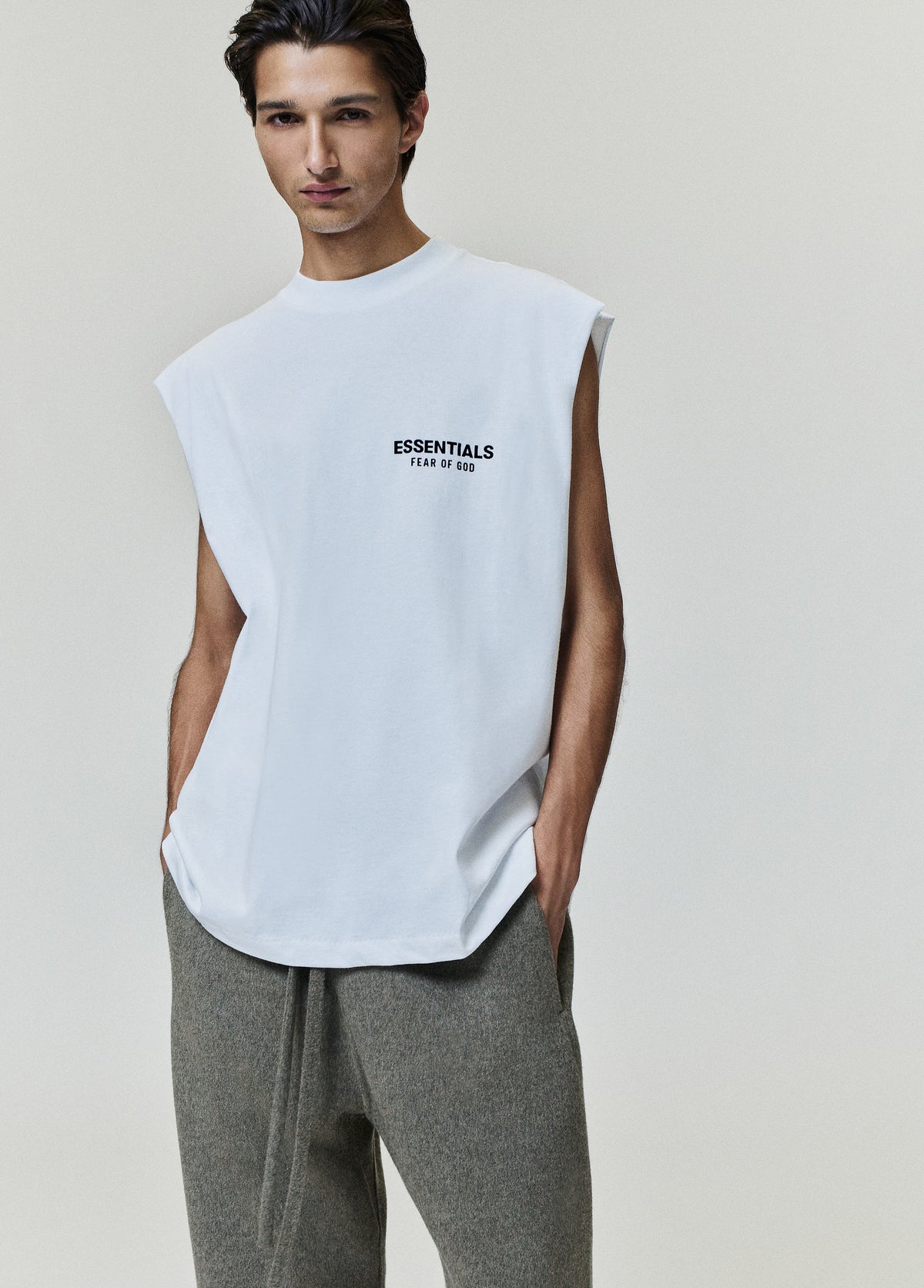 Essentials FW26 Classic Cut-Off white sleeveless top with raw armholes and chest Fear of God logo worn with gray trousers in a studio Tops view