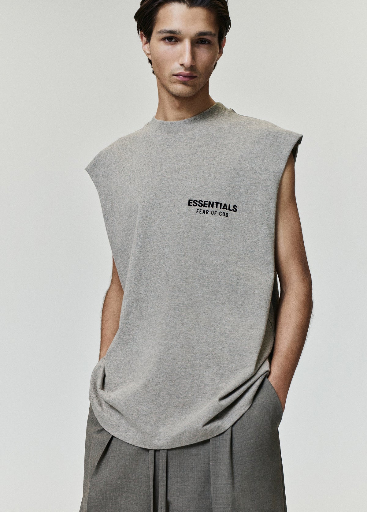 Essentials FW26 Classic Cut-Off gray sleeveless top with raw armholes and Fear of God logo worn by a model with tailored gray trousers in a studio Tops view