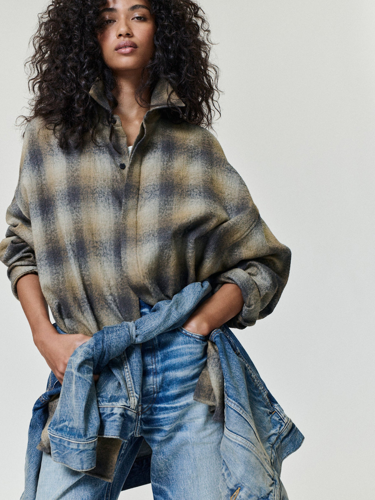 Woman with voluminous curly hair wearing an oversized plaid zip jacket with rolled sleeves and blue jeans with a denim jacket tied at the waist against a pale gray background Tops Bottoms Outerwear