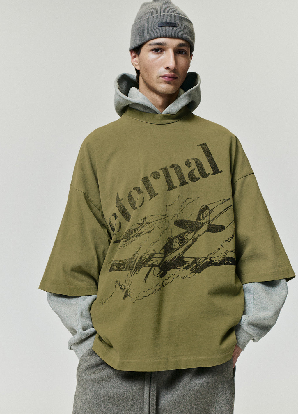 Flight Tee in olive green layered over a gray hoodie with oversized Eternal lettering and a vintage aircraft graphic worn by a model as Tops