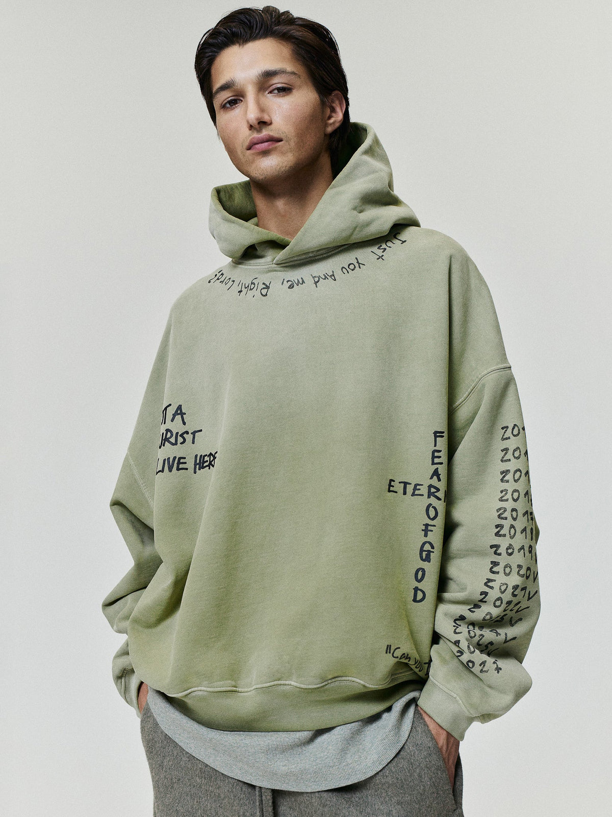 Hand Written Hoodie in sage green with handwritten Fear of God graphics on the hood and sleeves, styled on a model with gray layering as Tops