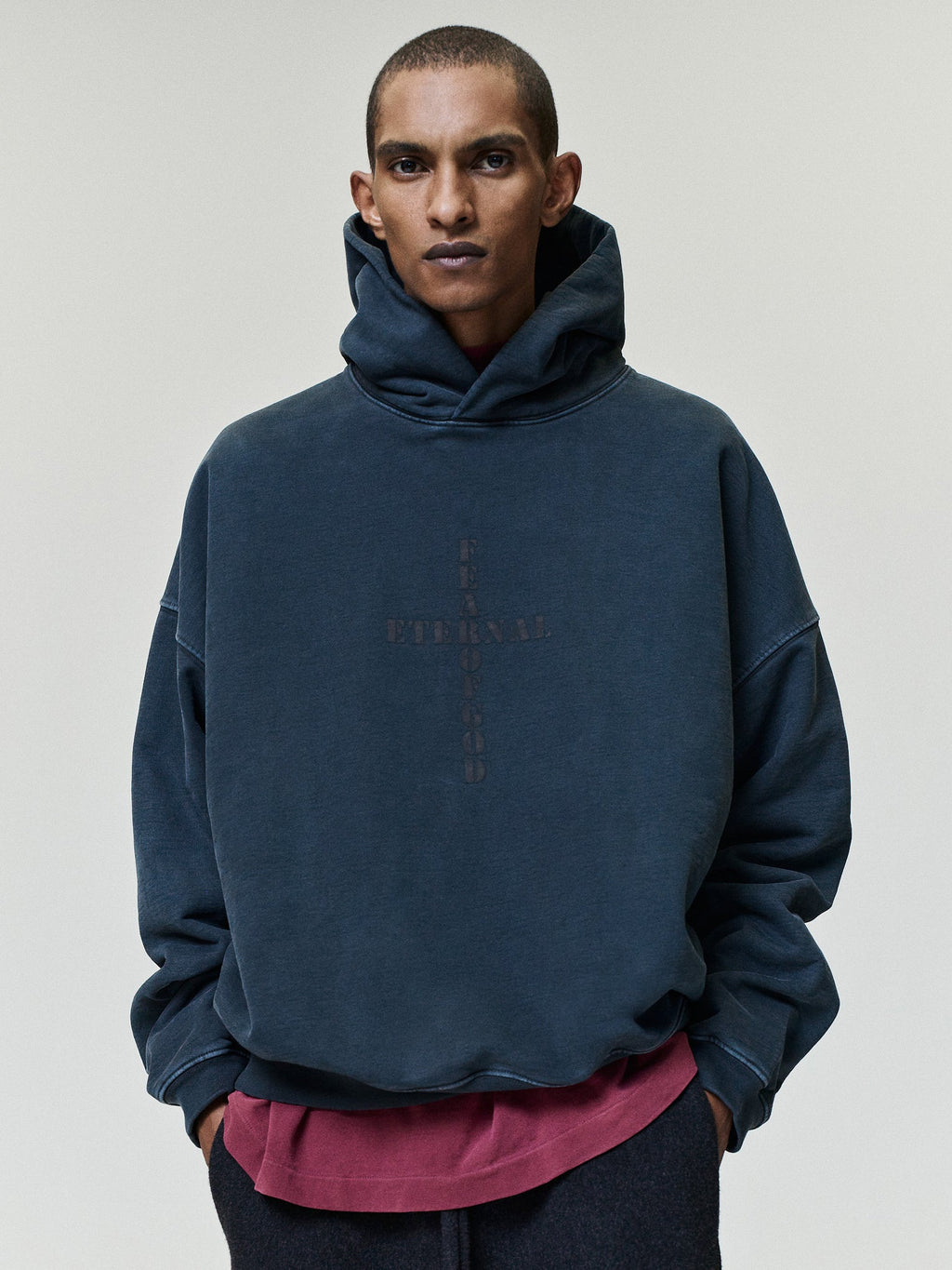 Cross Hoodie shown on a model in dark navy with an oversized fit layered hood long sleeves and subtle cross graphic over a burgundy shirt as Tops