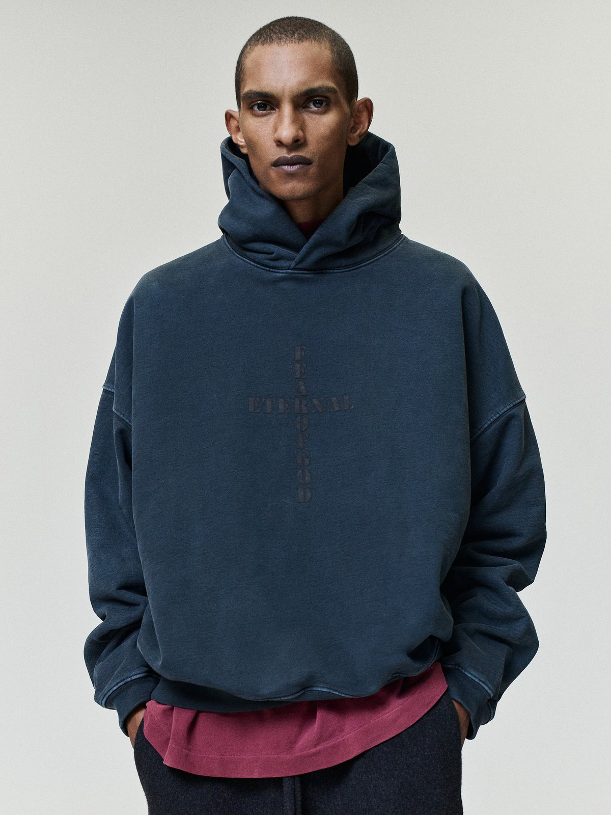 Cross Hoodie shown on a model in dark navy with an oversized fit layered hood long sleeves and subtle cross graphic over a burgundy shirt as Tops