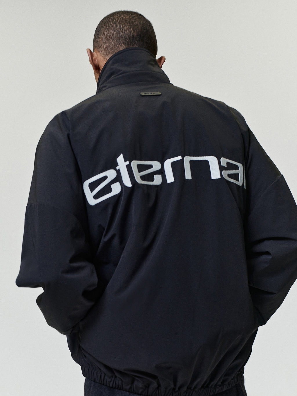 Bonded Nylon Tech Windbreaker in black shown from the back on a model with a high stand collar elastic hem and large Eternal graphic as Outerwear