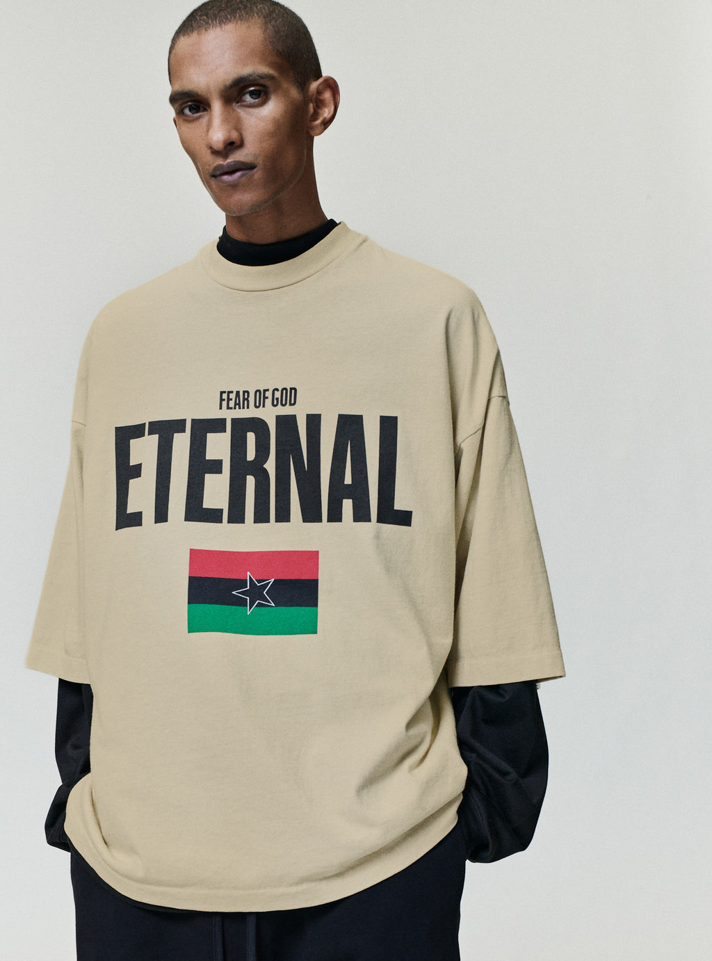Reflection Eternal Tee in beige with Fear of God Eternal lettering and red black green flag graphic with star worn over black sleeves as Tops