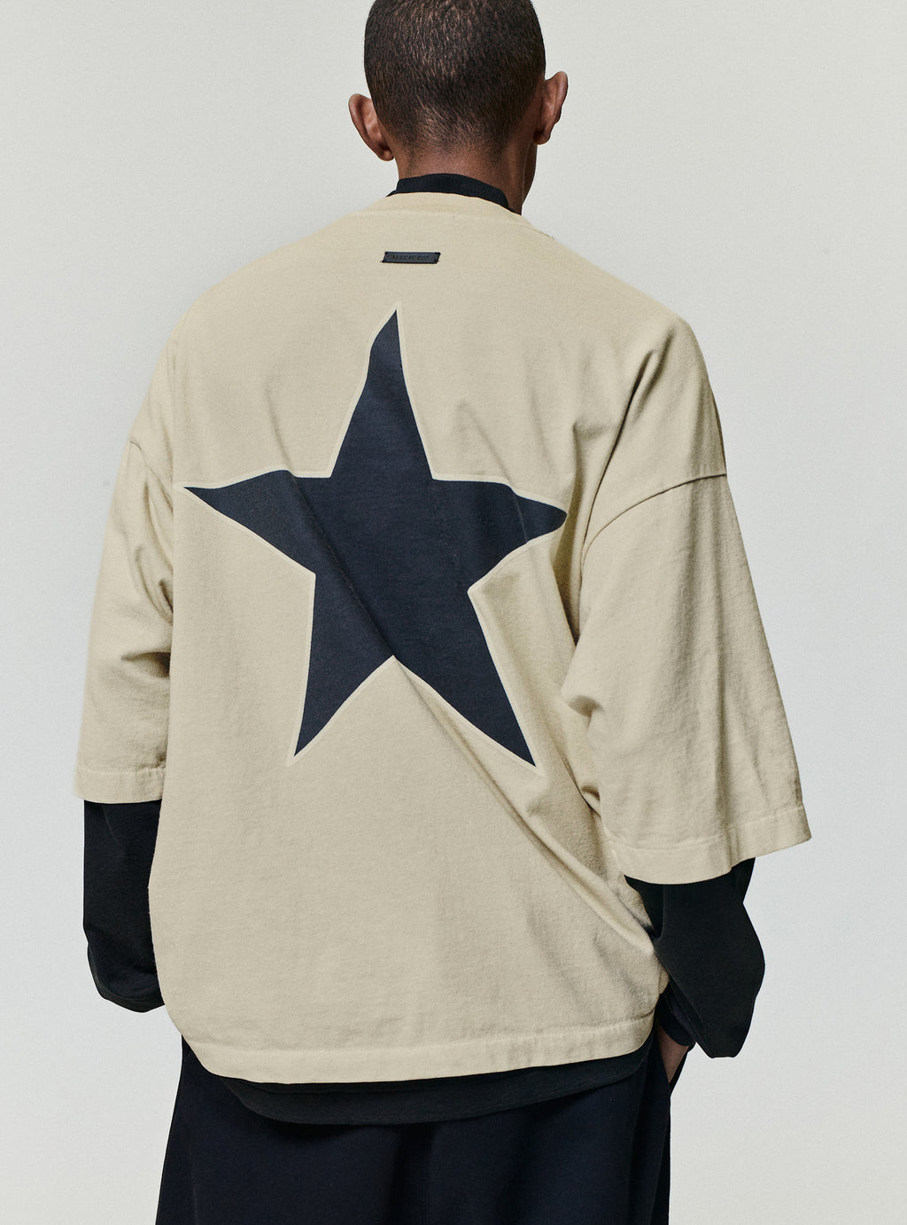 Reflection Eternal Tee in beige viewed from the back with a large dark star graphic and layered black long sleeves as Tops
