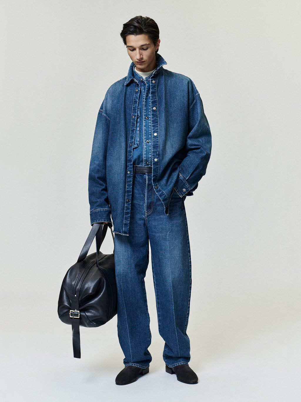 Male model wearing a dark denim jacket with matching wide leg jeans and black shoes while carrying a black leather bag against a pale studio background Outerwear Bottoms Footwear