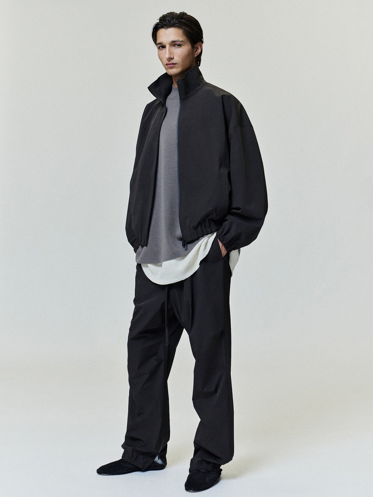 Bonded Nylon Track Jacket in black worn by a model with a high stand collar elastic hem and gathered sleeves layered over a gray top as Outerwear