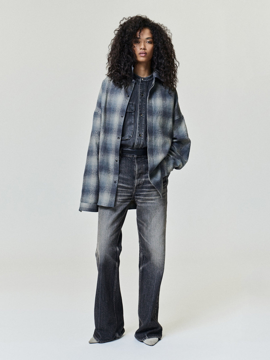 Woman with curly hair wearing a blue gray plaid overshirt over a dark denim shirt and gray wide leg jeans in a light gray studio Outerwear Tops Bottoms