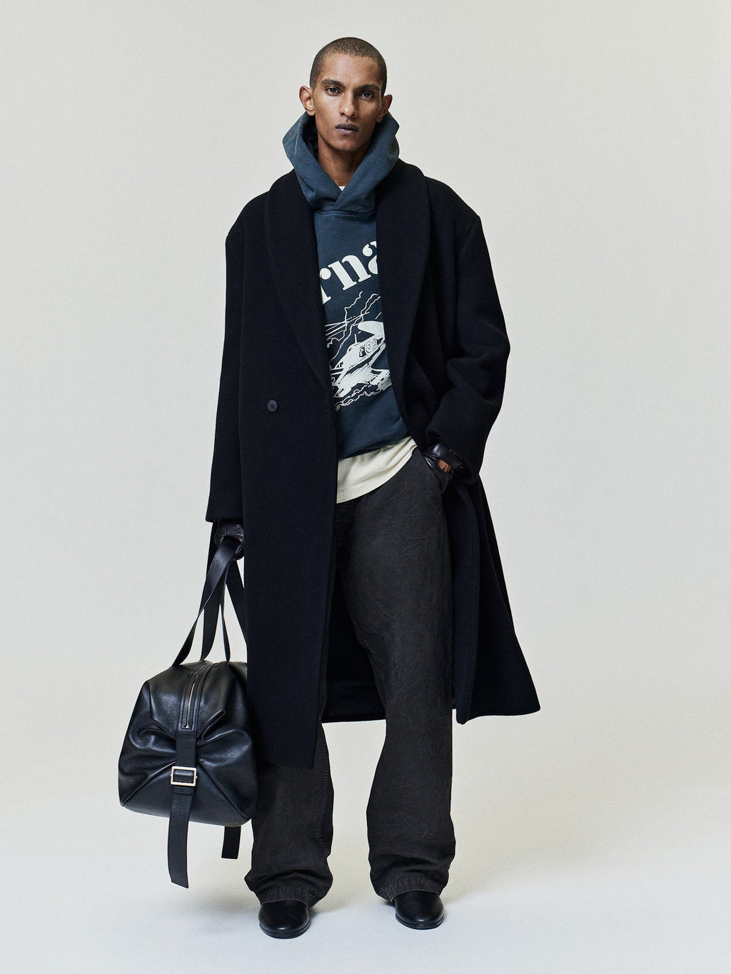 Flight Hoodie in dark blue with vintage aircraft artwork styled on a model beneath a black overcoat with relaxed trousers and leather bag as Tops