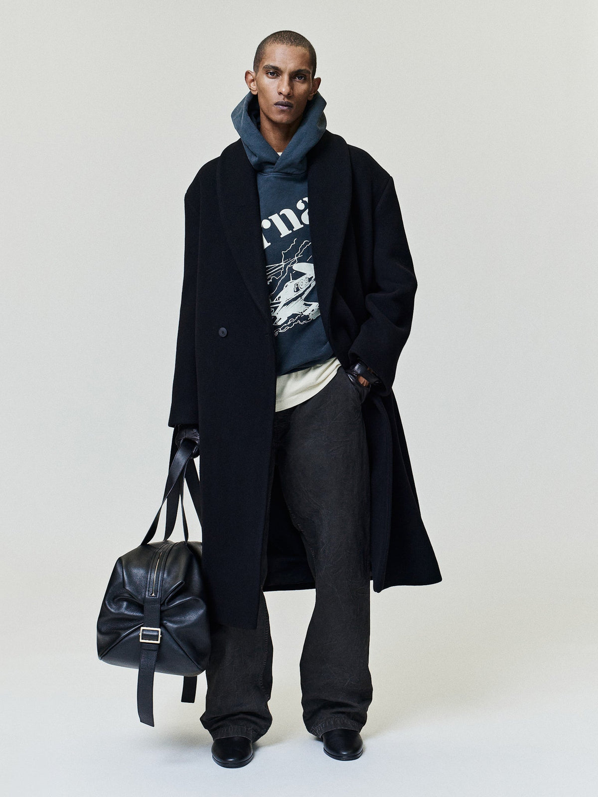 Flight Hoodie in dark blue with vintage aircraft artwork styled on a model beneath a black overcoat with relaxed trousers and leather bag as Tops