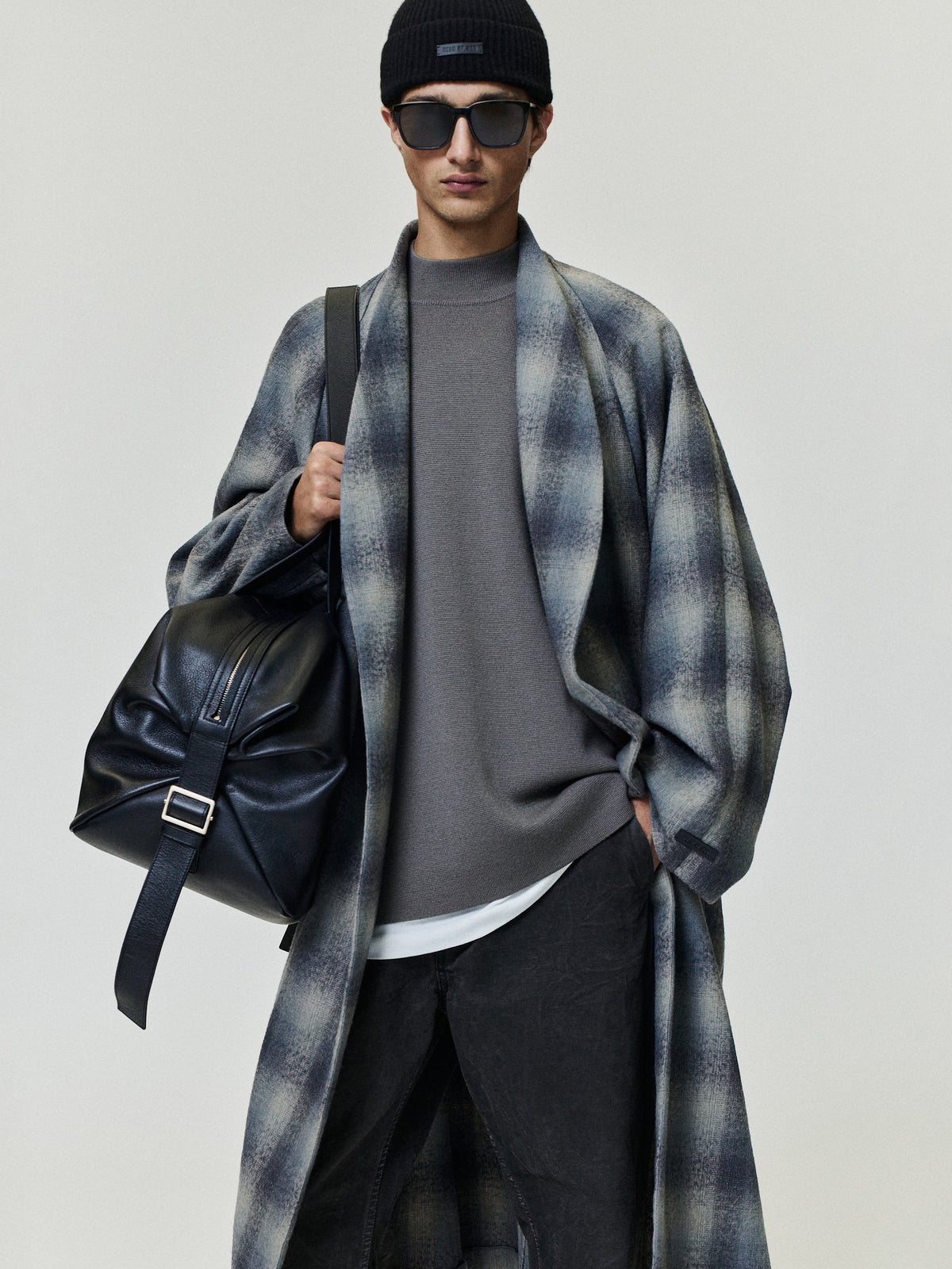Man in a black beanie and sunglasses wearing a long gray plaid overcoat over a charcoal sweater and white shirt with dark trousers and a leather shoulder bag Outerwear Tops Bottoms
