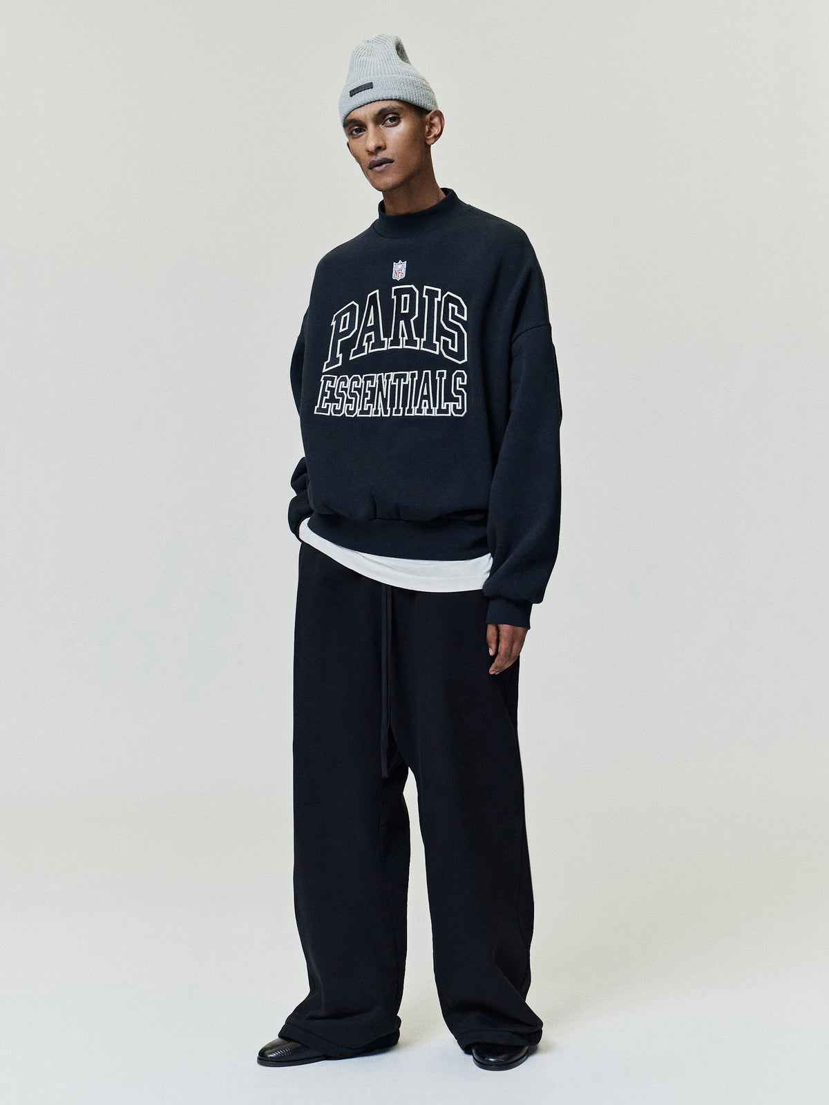 NFL Paris Boxy Crewneck in dark navy with white Paris Essentials lettering styled with relaxed trousers and a gray beanie against a plain backdrop Tops