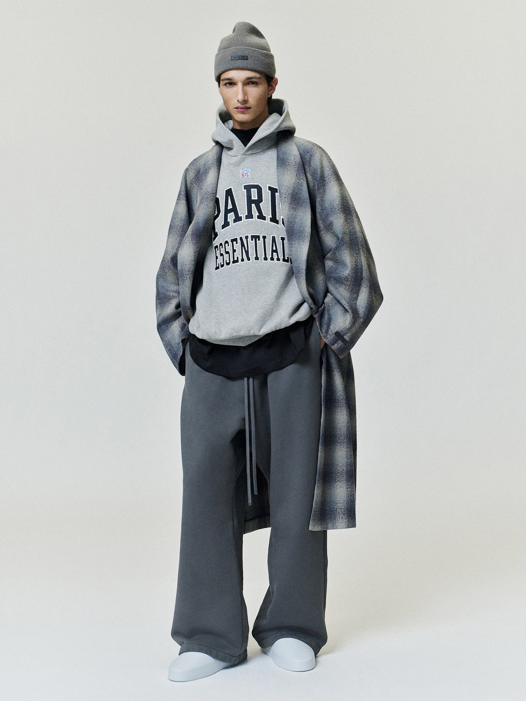 NFL Paris 90s Relaxed Hoodie in gray with Paris Essentials lettering worn under a long plaid coat with wide leg trousers beanie and white shoes Tops