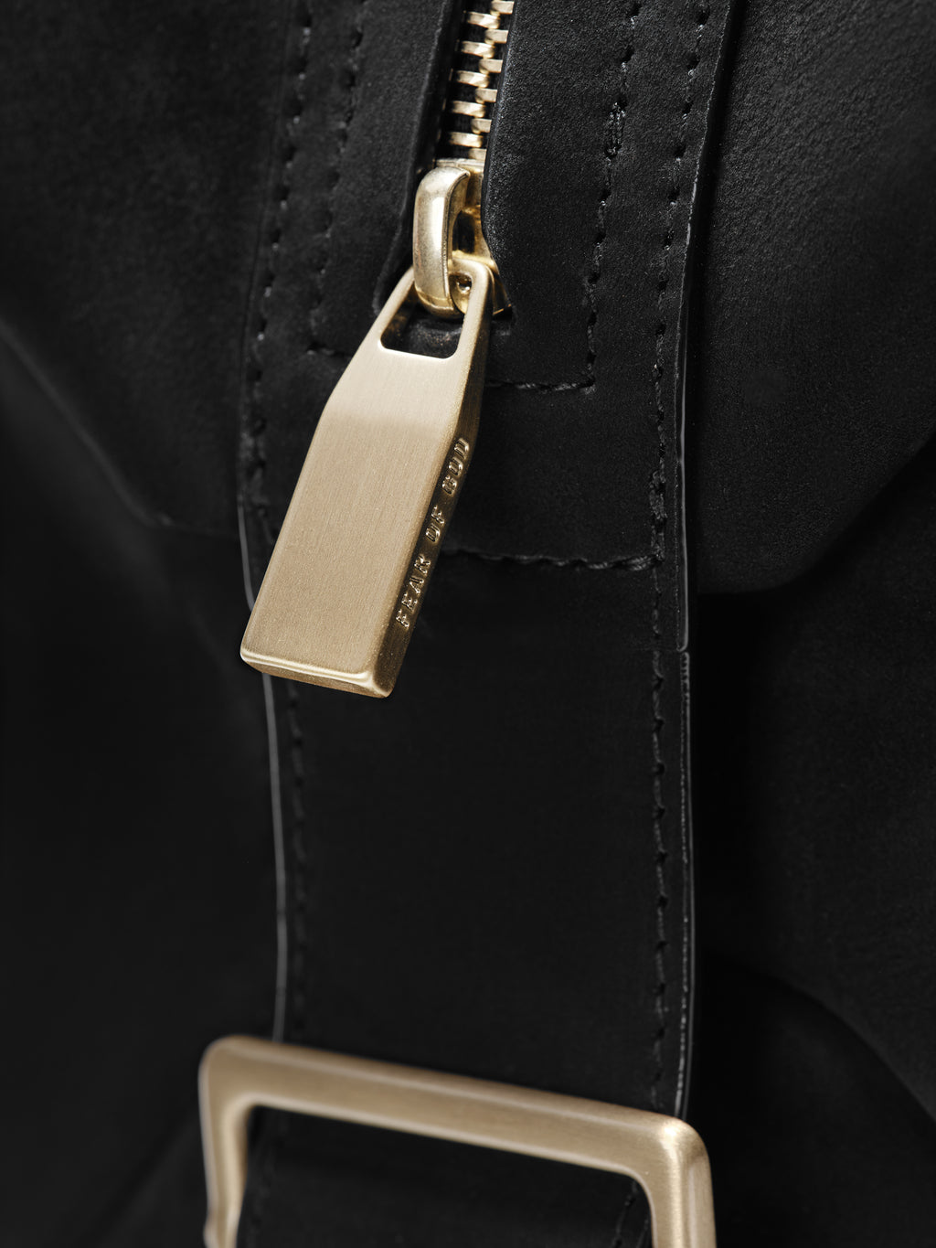 Close-up of a matte black Large Belted Duffle showcasing a smooth leather strap with precise stitching, a brushed gold-tone zipper pull, and a matching rectangular buckle.