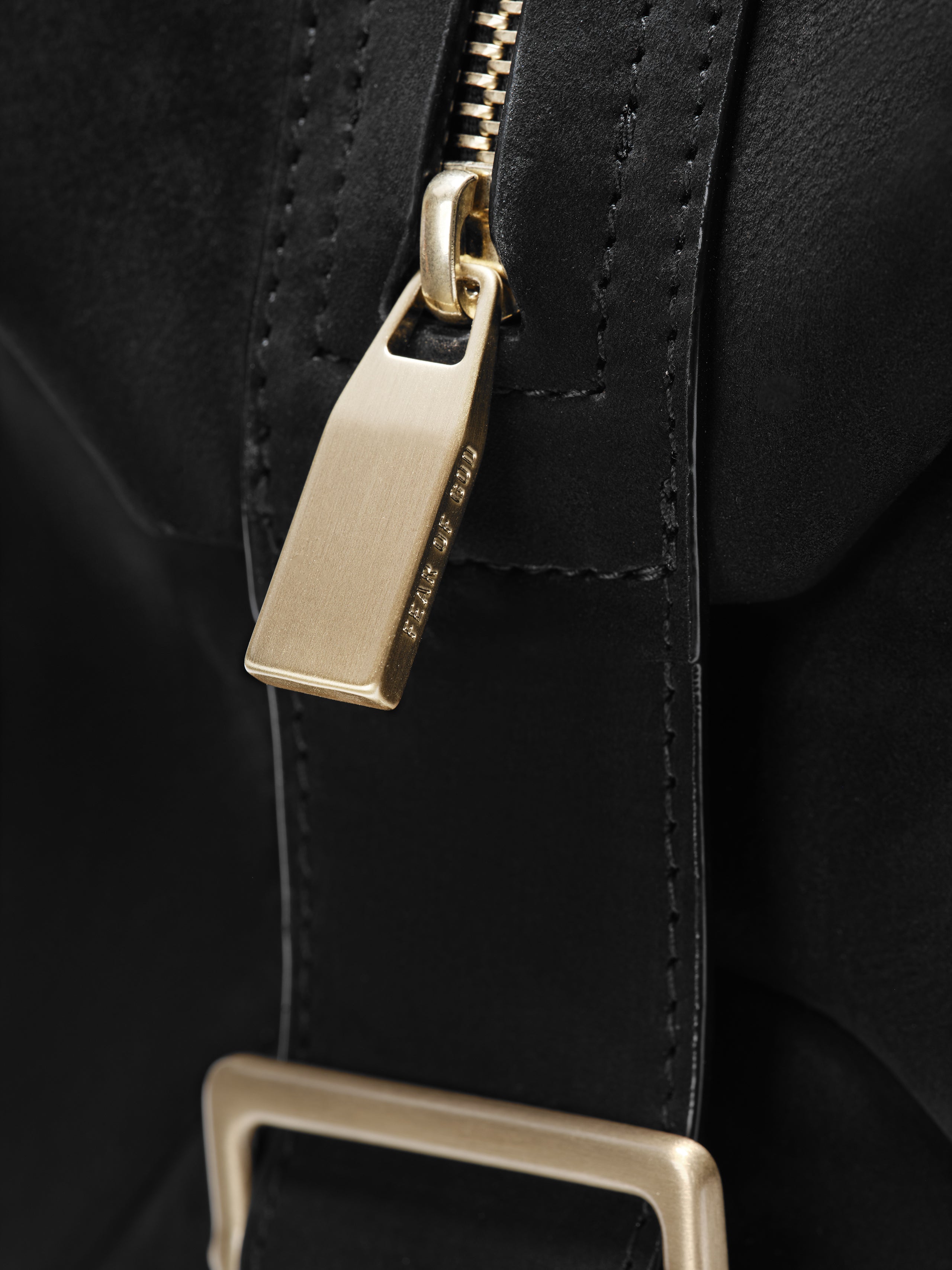 Close-up of a matte black Large Belted Duffle showcasing a smooth leather strap with precise stitching, a brushed gold-tone zipper pull, and a matching rectangular buckle.