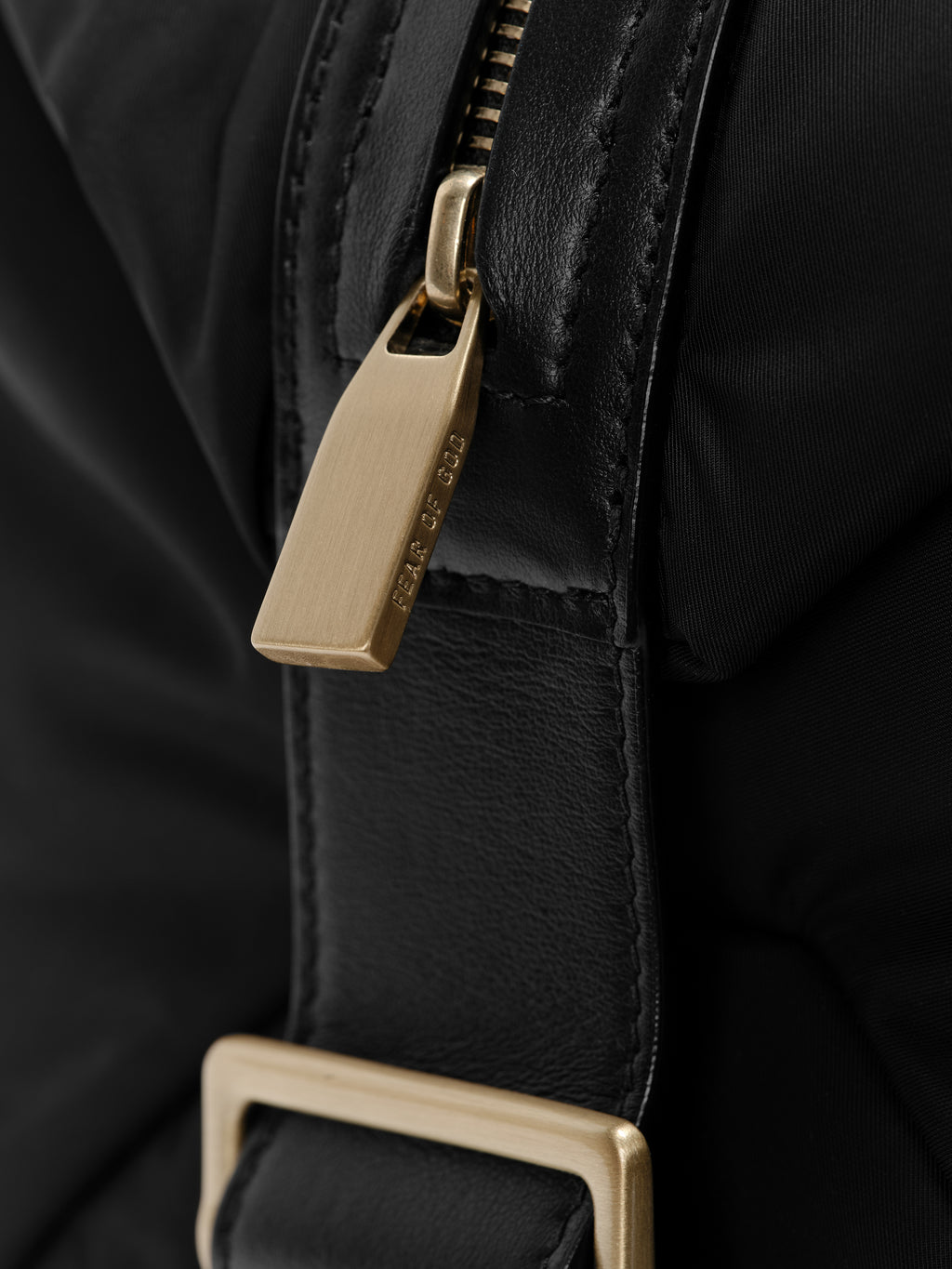 Close-up of a black leather-trimmed zipper pull with engraved text on a textured black nylon bag strap detail