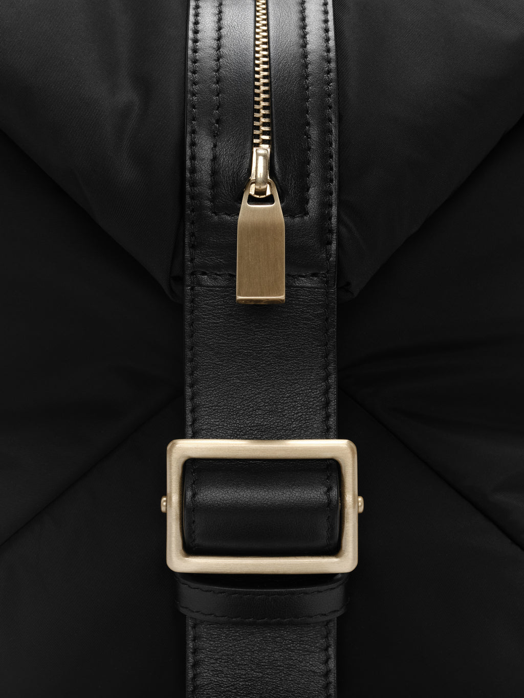 Close-up of gold zipper and leather strap with rectangular gold buckle on black nylon fabric background