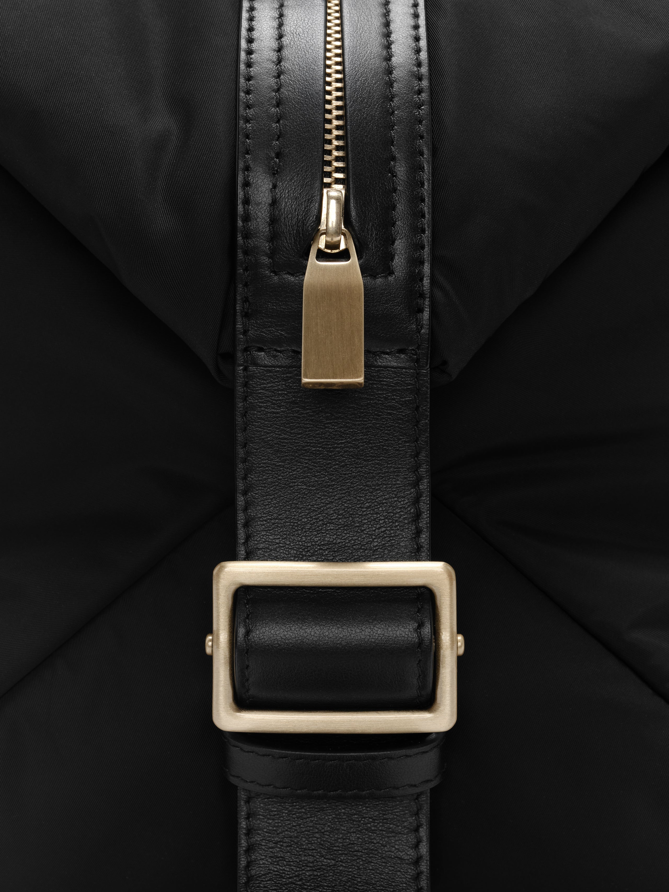 Close-up of gold zipper and leather strap with rectangular gold buckle on black nylon fabric background
