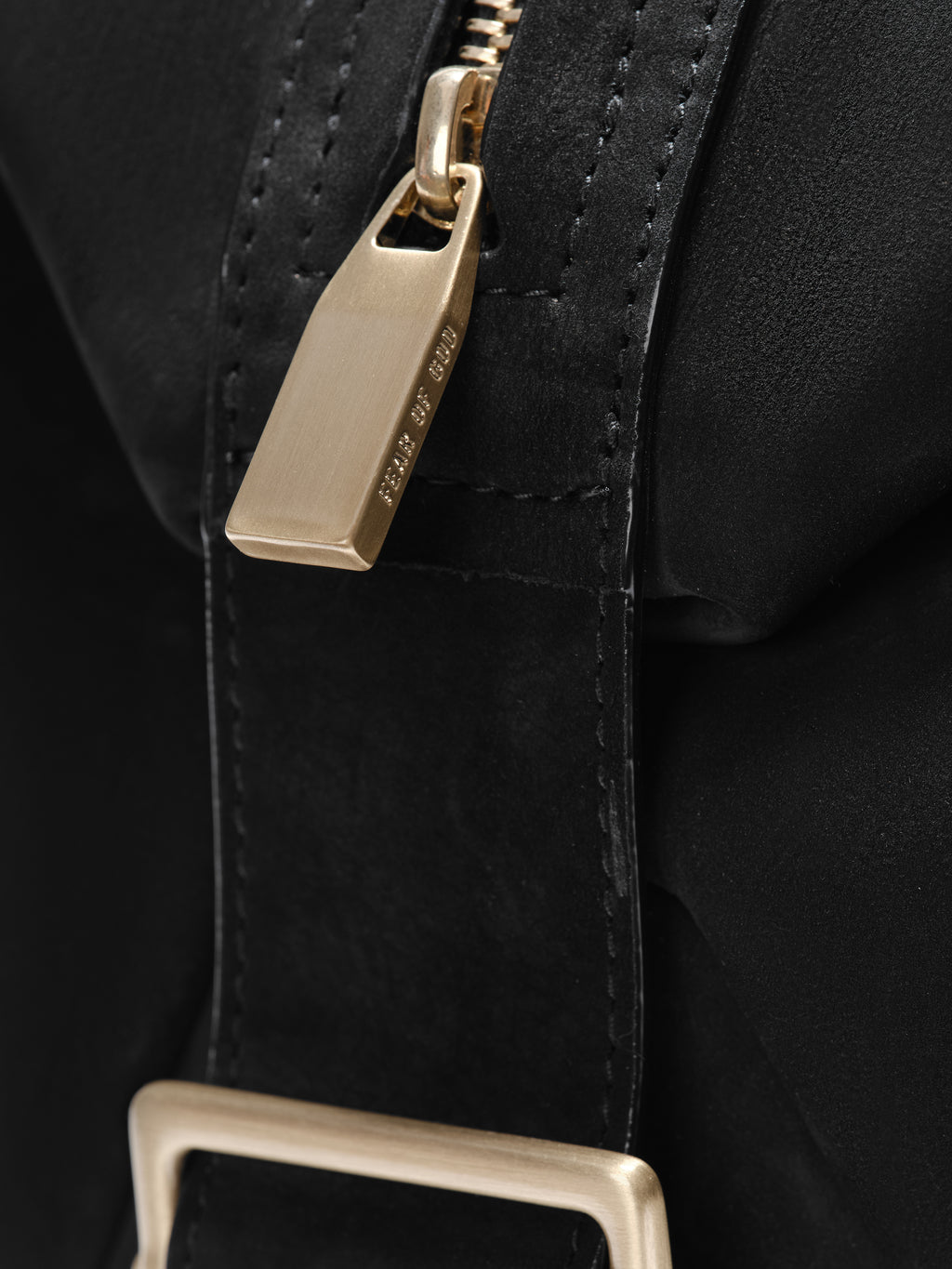 Close-up of the Small Belted Duffle in black highlighting a matte gold zipper pull engraved with subtle branding, contrasted against smooth black leather with detailed stitching.