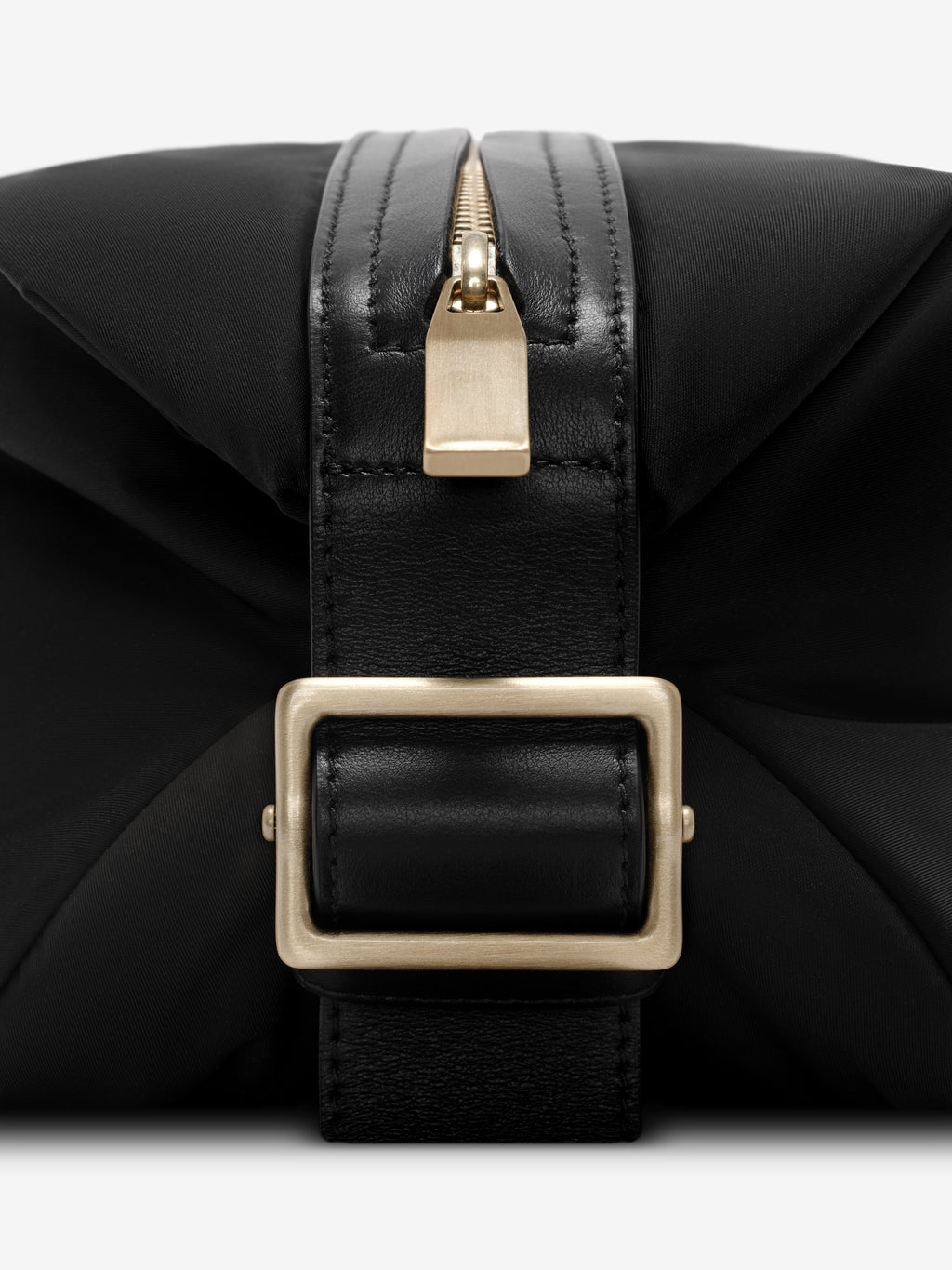 Close-up of gold zipper pull and matte gold rectangular buckle on black nylon pouch with leather strap detail