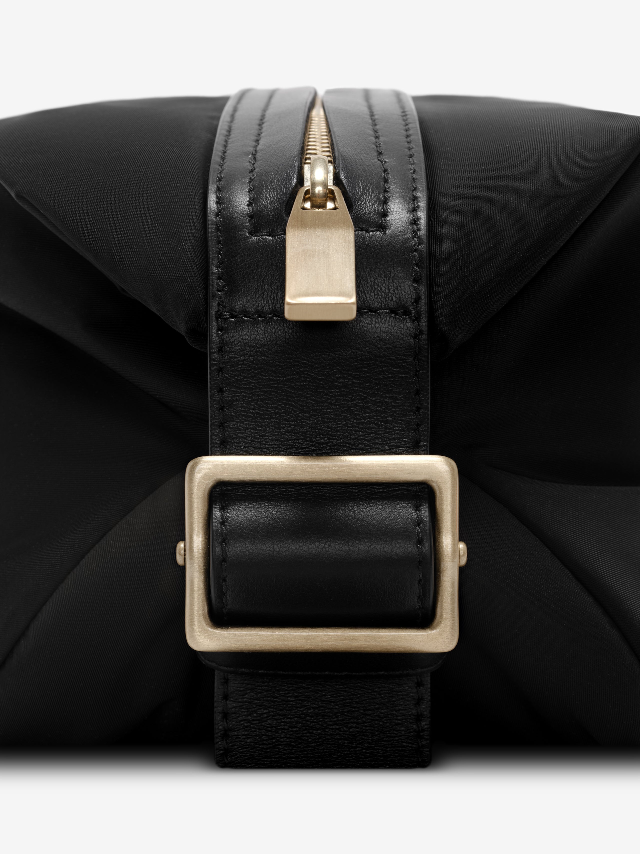 Close-up of gold zipper pull and matte gold rectangular buckle on black nylon pouch with leather strap detail