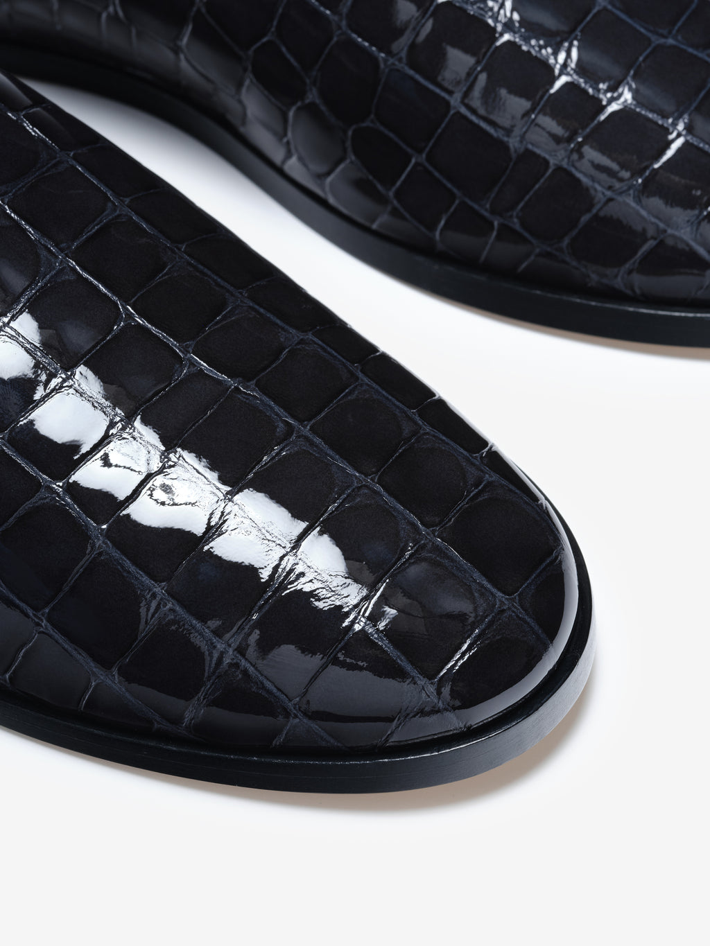 Close-up of shiny black shoes with embossed crocodile pattern and rounded toe on white background