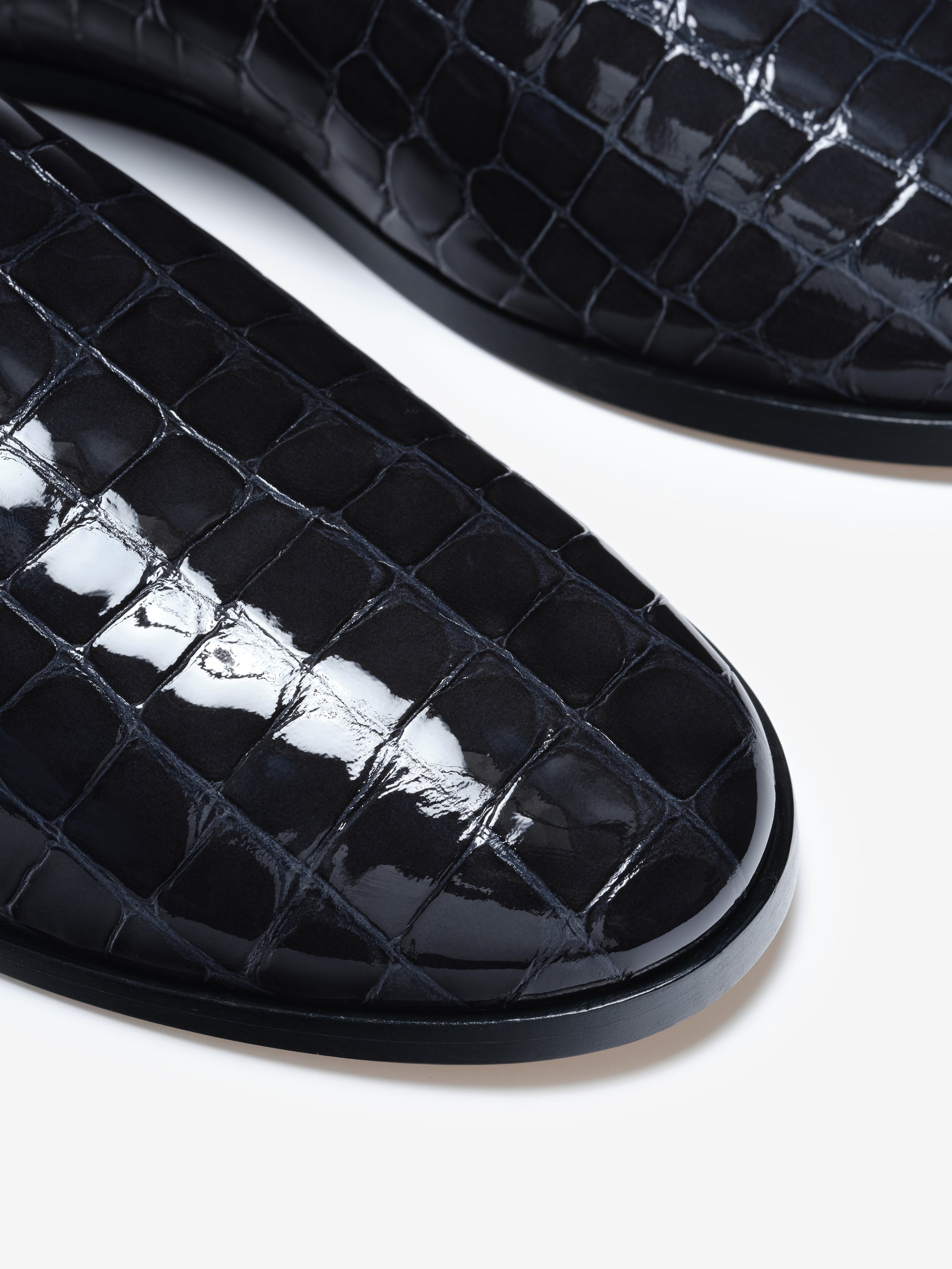 Close-up of shiny black shoes with embossed crocodile pattern and rounded toe on white background