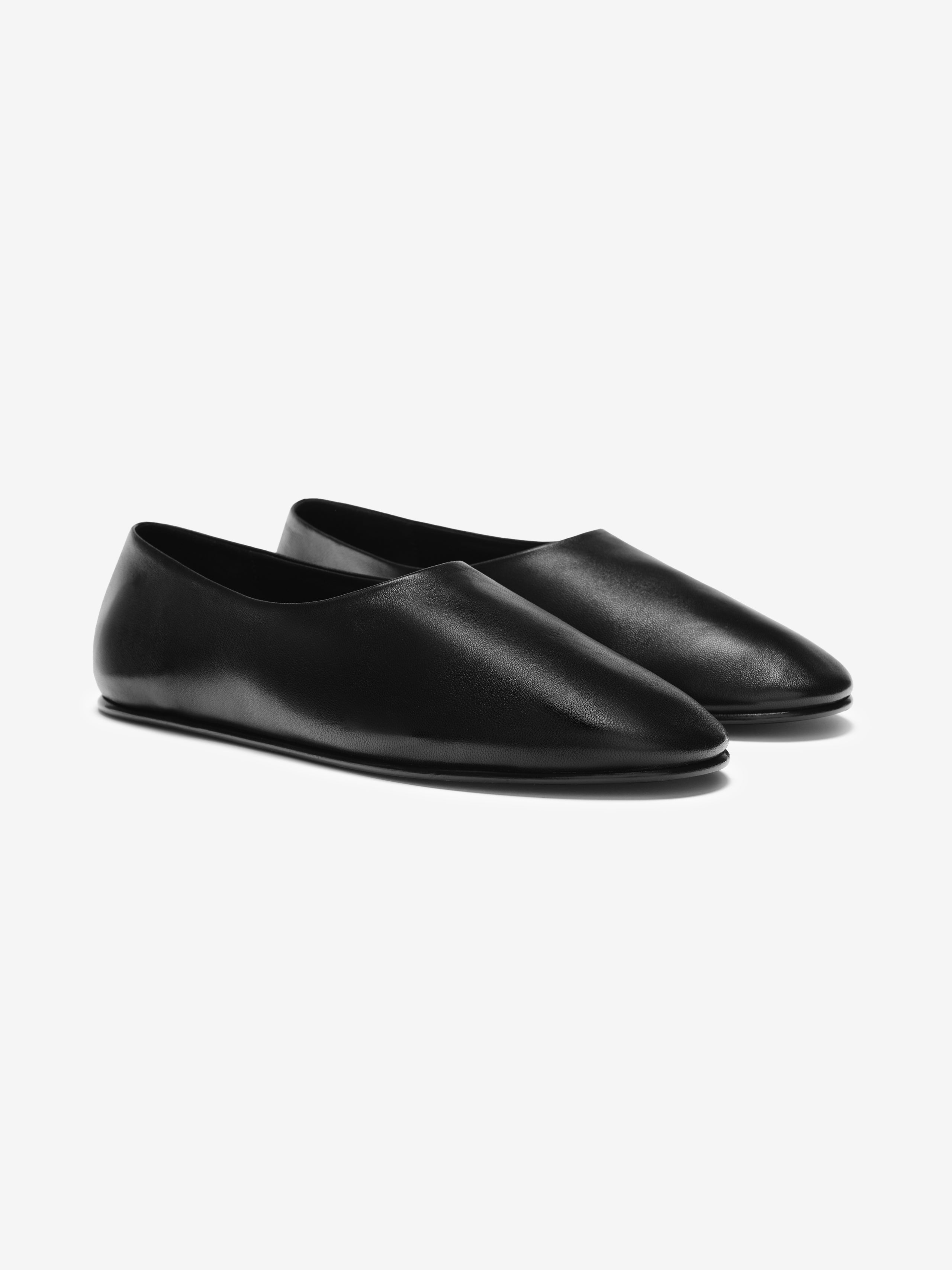 Pair of minimalist matte black leather slip-on shoes with rounded toe and flat sole on light gray background