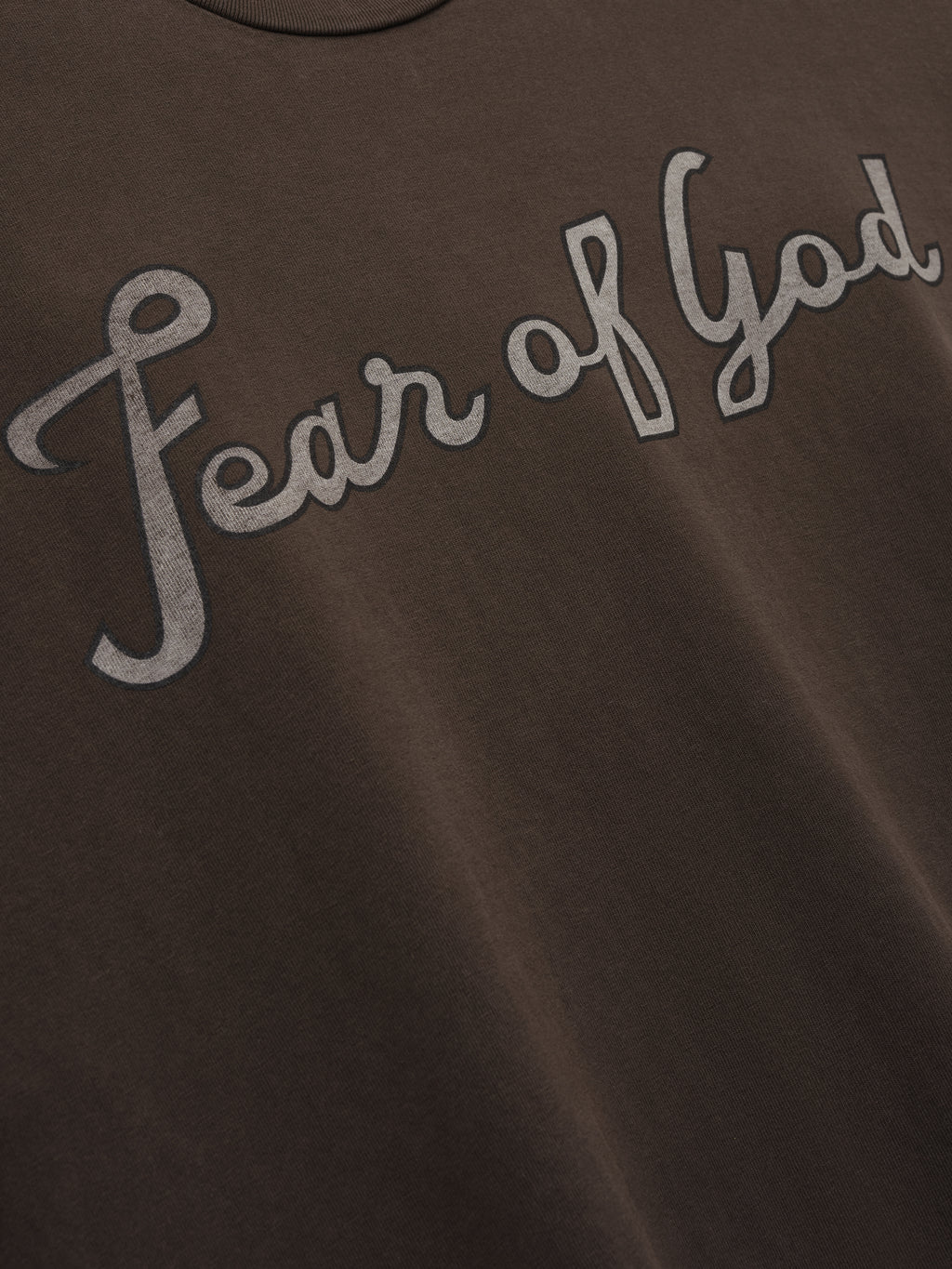 Close-up of dark brown fabric with "Fear of God" printed in large gray cursive letters across the chest