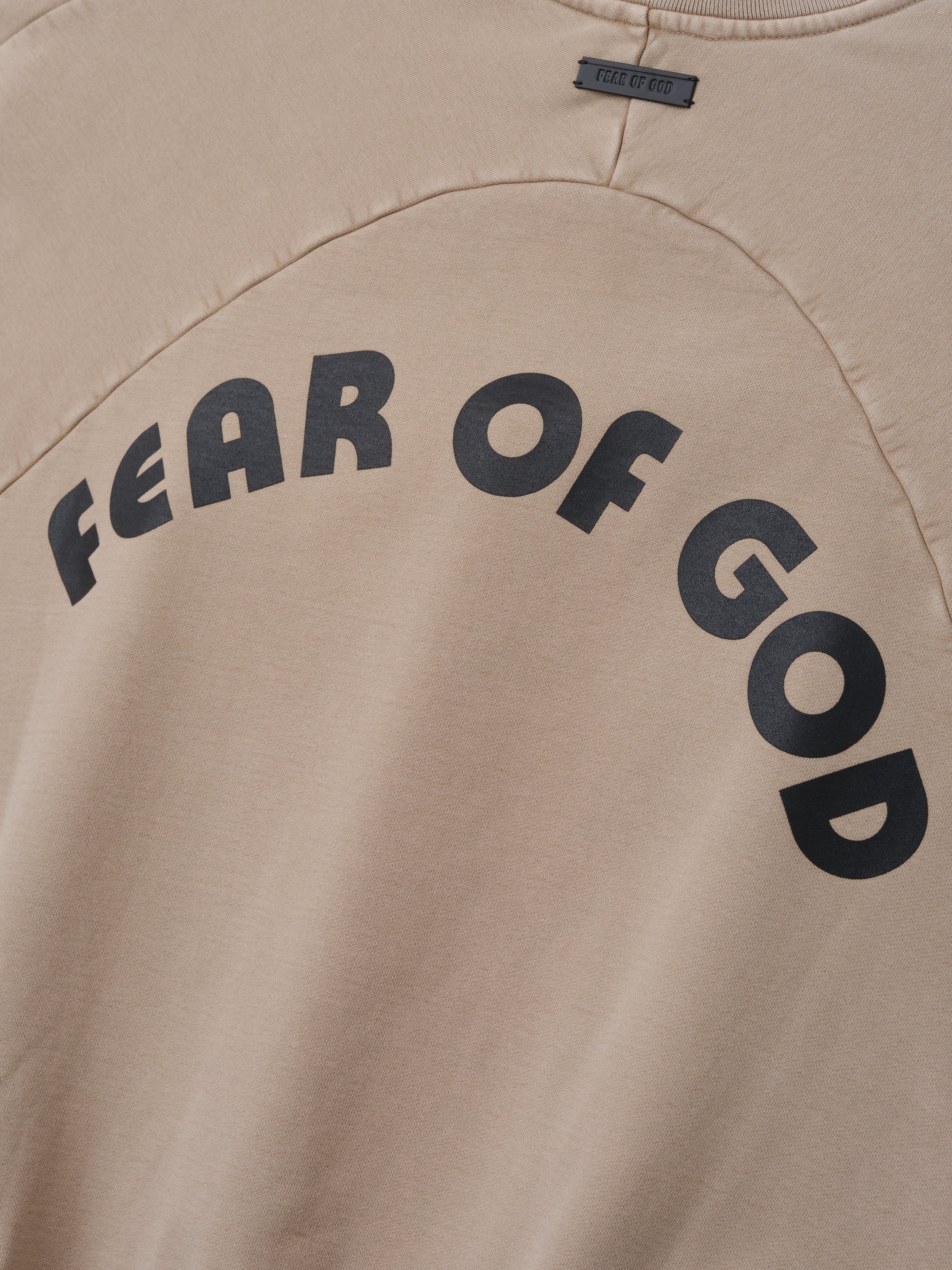Close-up of dune-toned cotton fabric with large black curved "FEAR OF GOD" text across the back panel