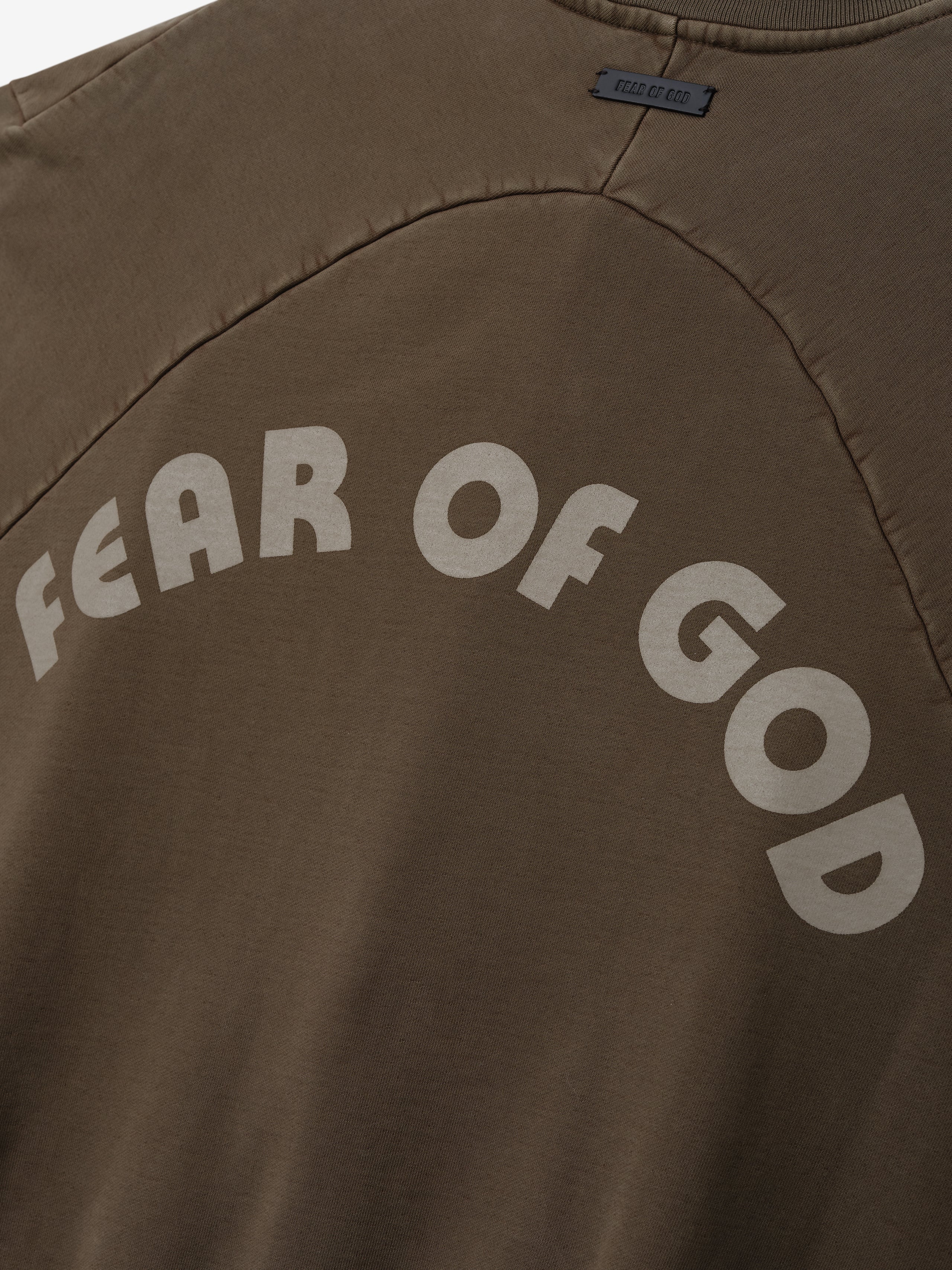 Close-up of faded brown cotton sweatshirt with light beige arched "FEAR OF GOD" text on back panel and visible stitching detail