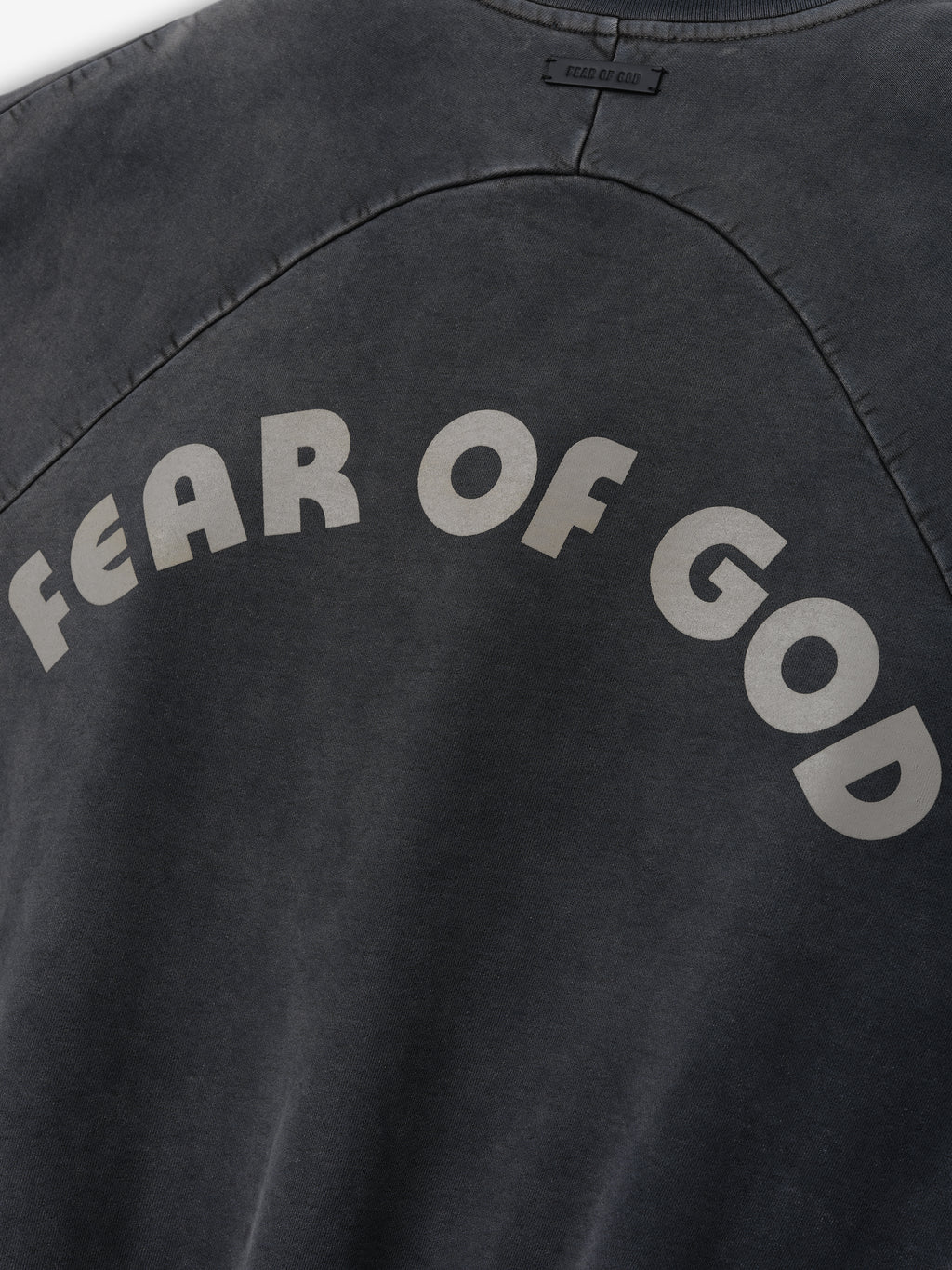 Close-up of faded black cotton sweatshirt with light gray arched "FEAR OF GOD" text on back panel