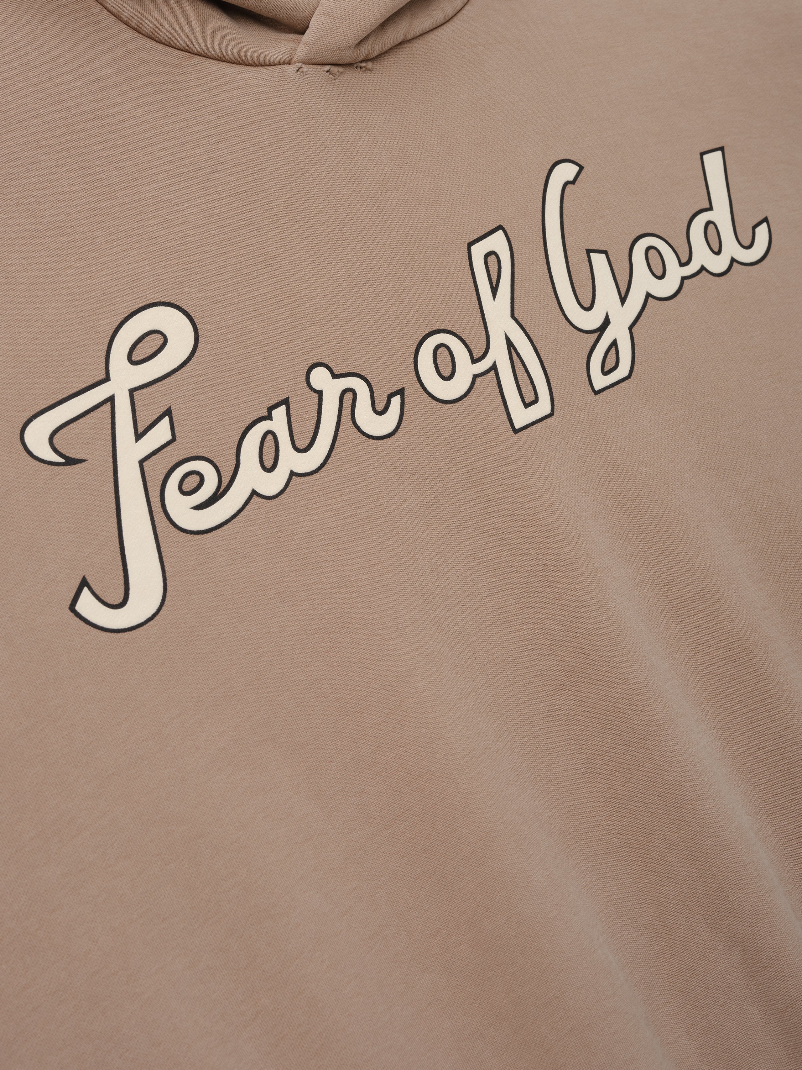 Close-up of a dune-colored hoodie with cream "Fear of God" cursive script printed diagonally across the chest