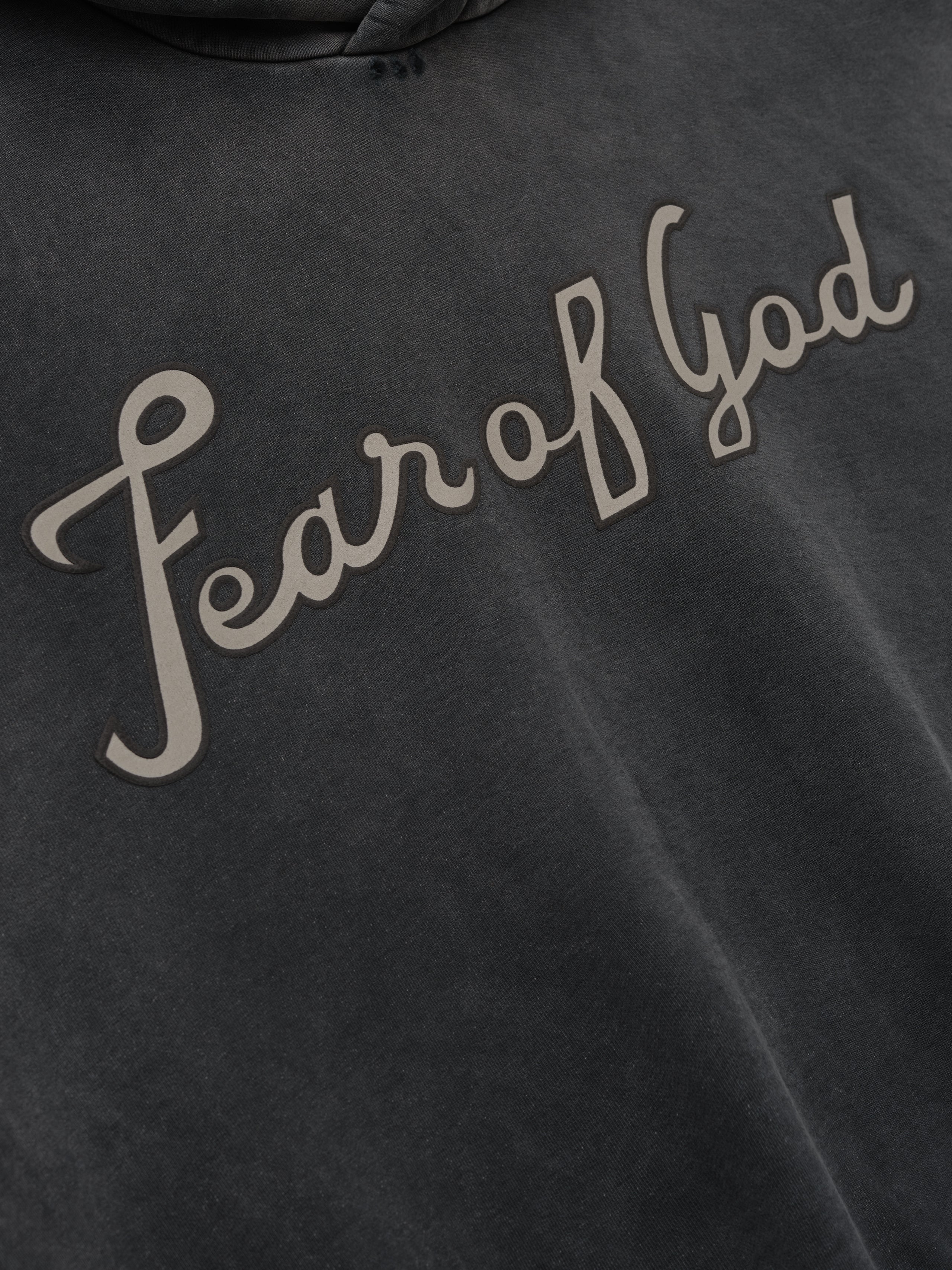 Close-up of off-black washed cotton fabric with raised beige cursive "Fear of God" text across front panel