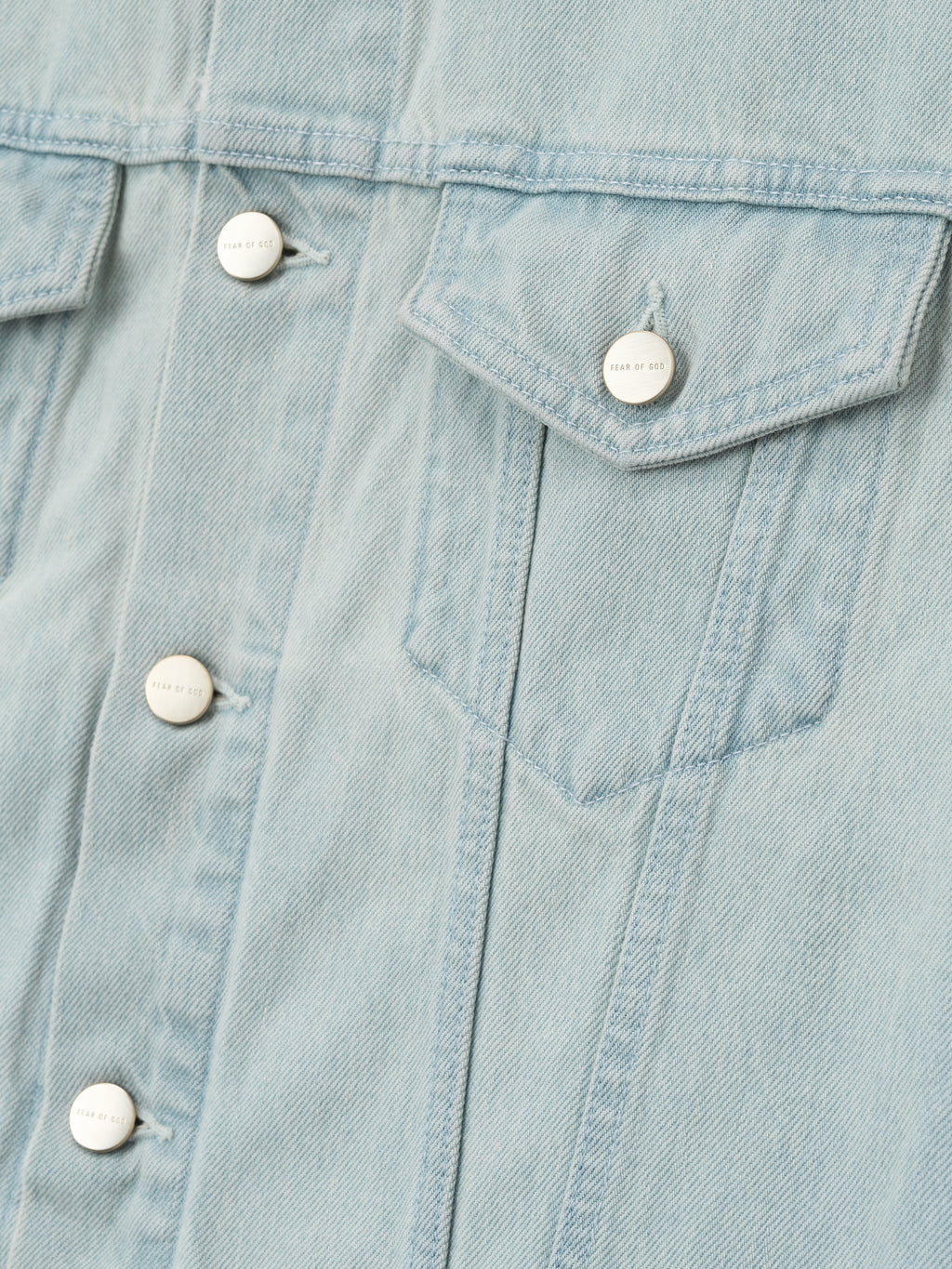 close-up of light bleached denim fabric with flap chest pocket and Fear of God metal shank buttons on jacket front panel