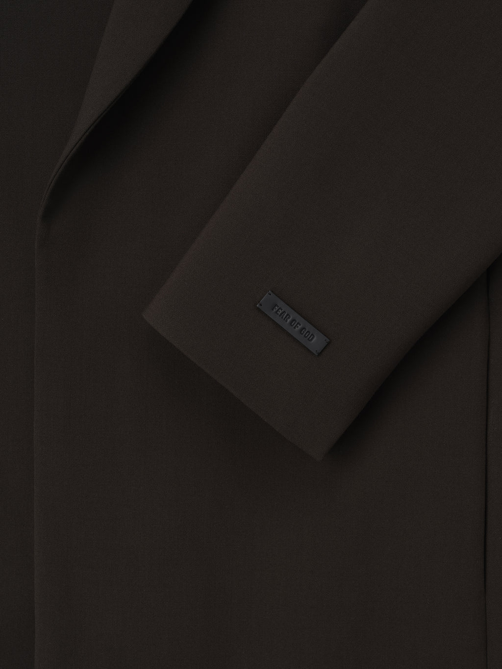 close-up of dark virgin wool crewneck jacket sleeve featuring matte Fear of God logo tag and smooth fabric texture
