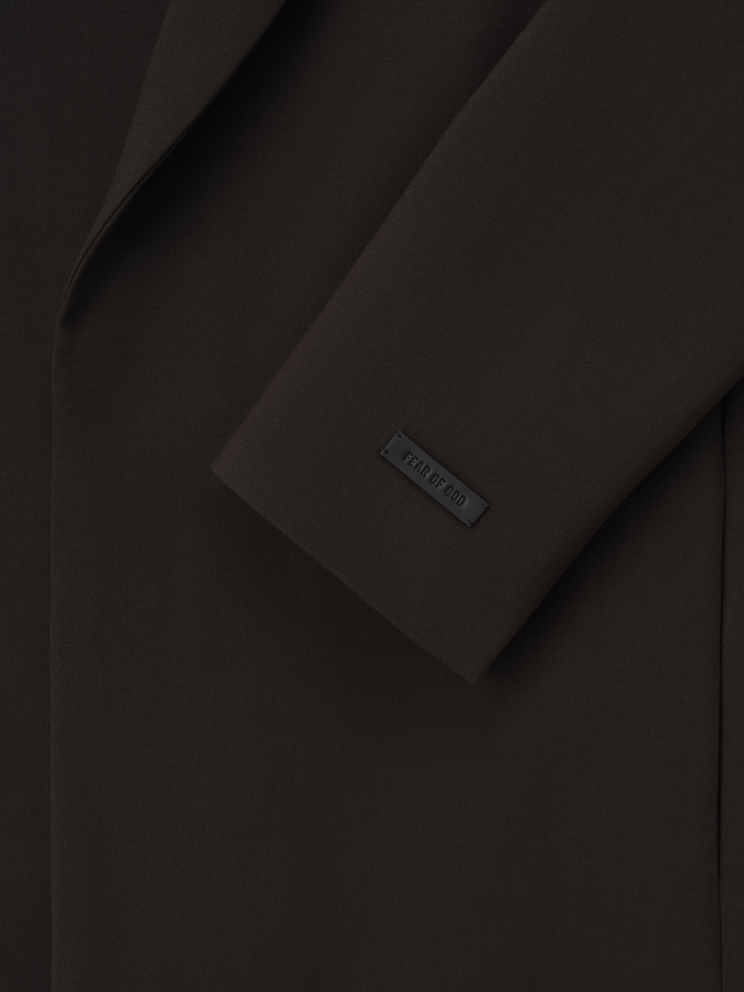 close-up of dark virgin wool crewneck jacket sleeve featuring matte Fear of God logo tag and smooth fabric texture