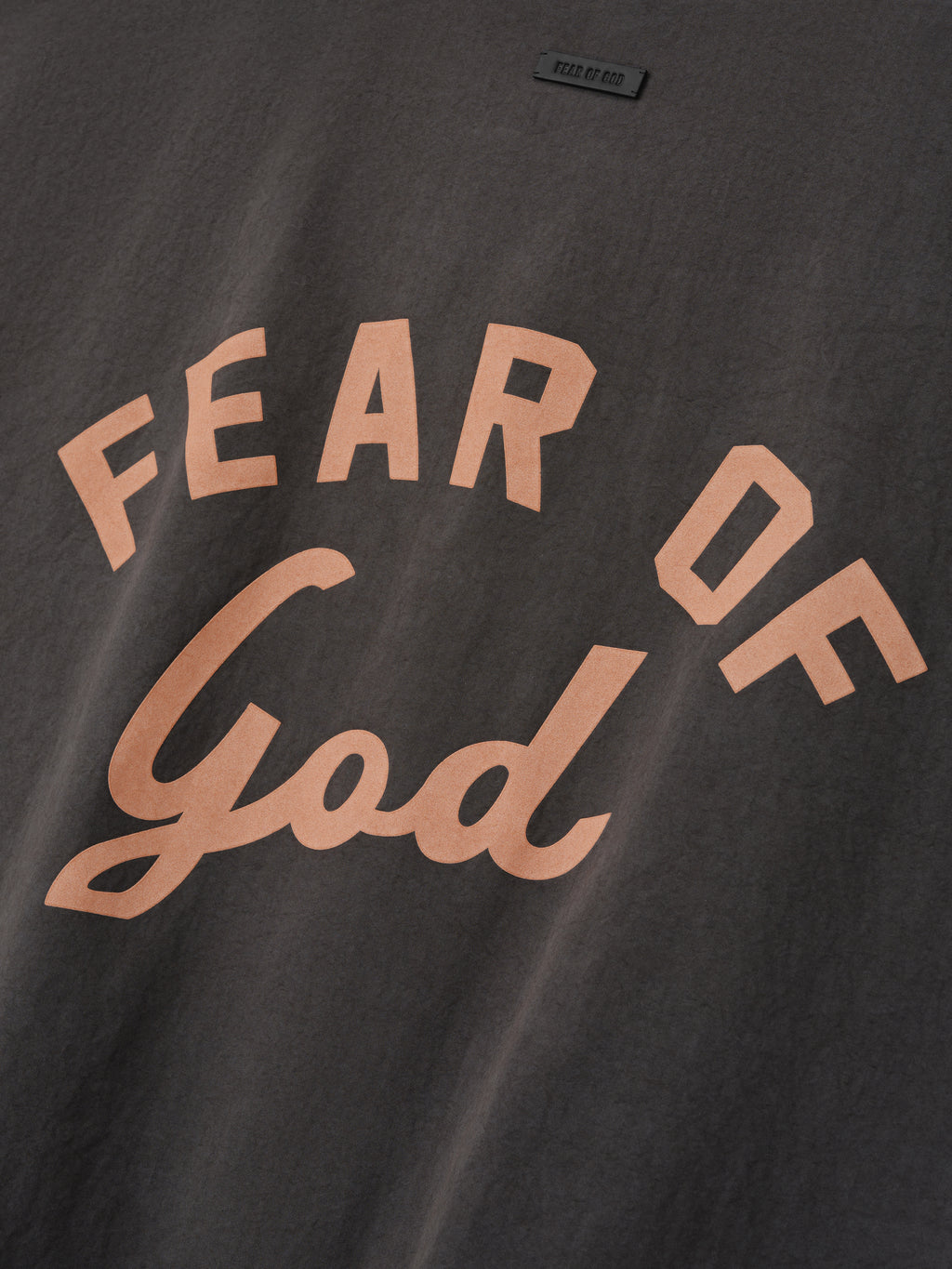 Close-up of dark gray fabric with large peach-colored "FEAR OF God" text in block and script fonts