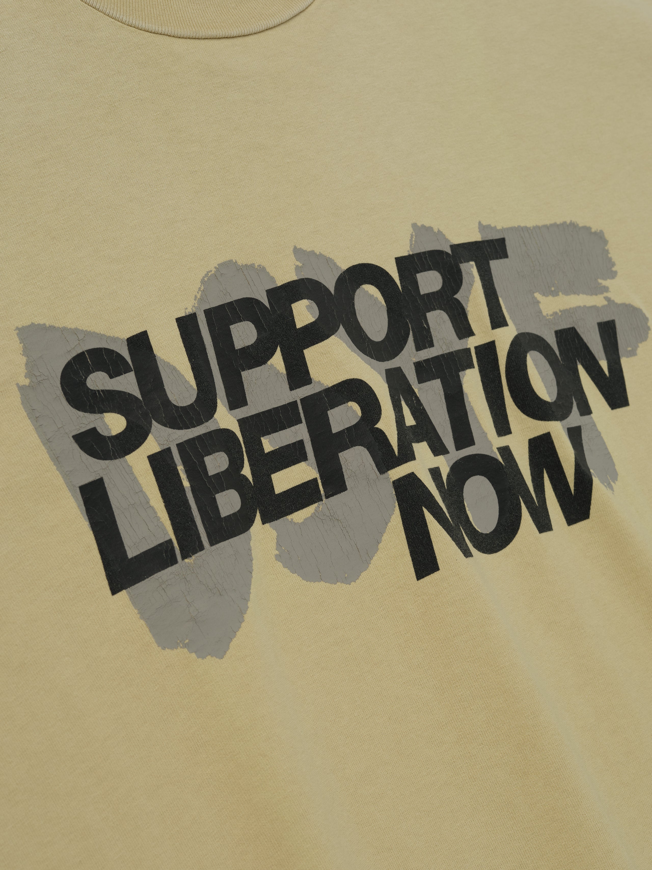 Close-up of clay Liberation Now Tee showing black "Support Liberation Now" text over gray brushstroke background centered on fabric