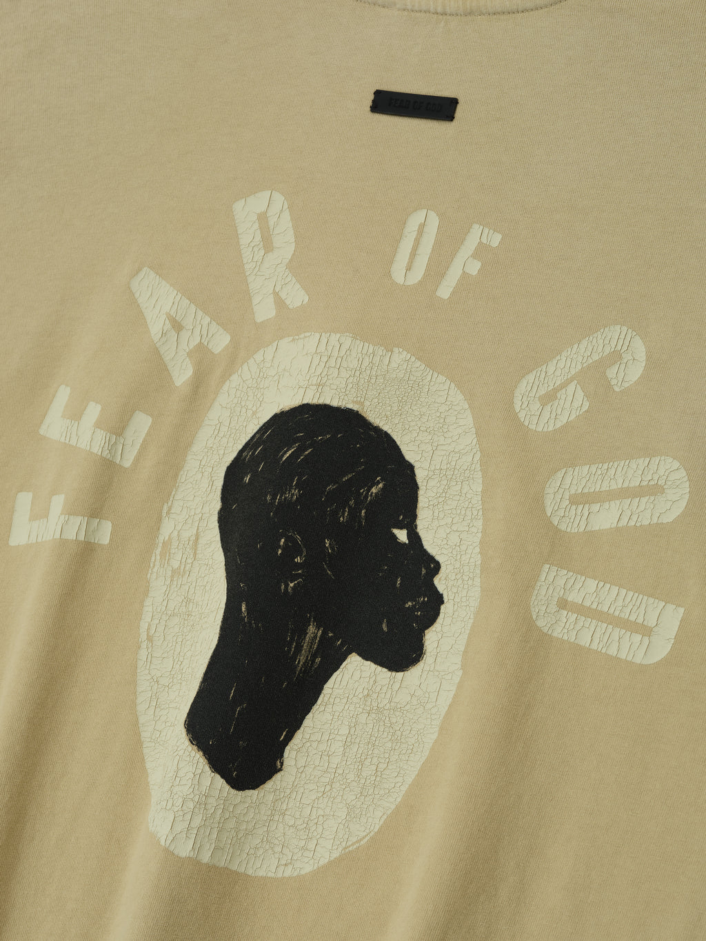 Close-up of clay Liberation Now Tee showing distressed black profile silhouette and cracked pale text "FEAR OF GOD" on chest fabric